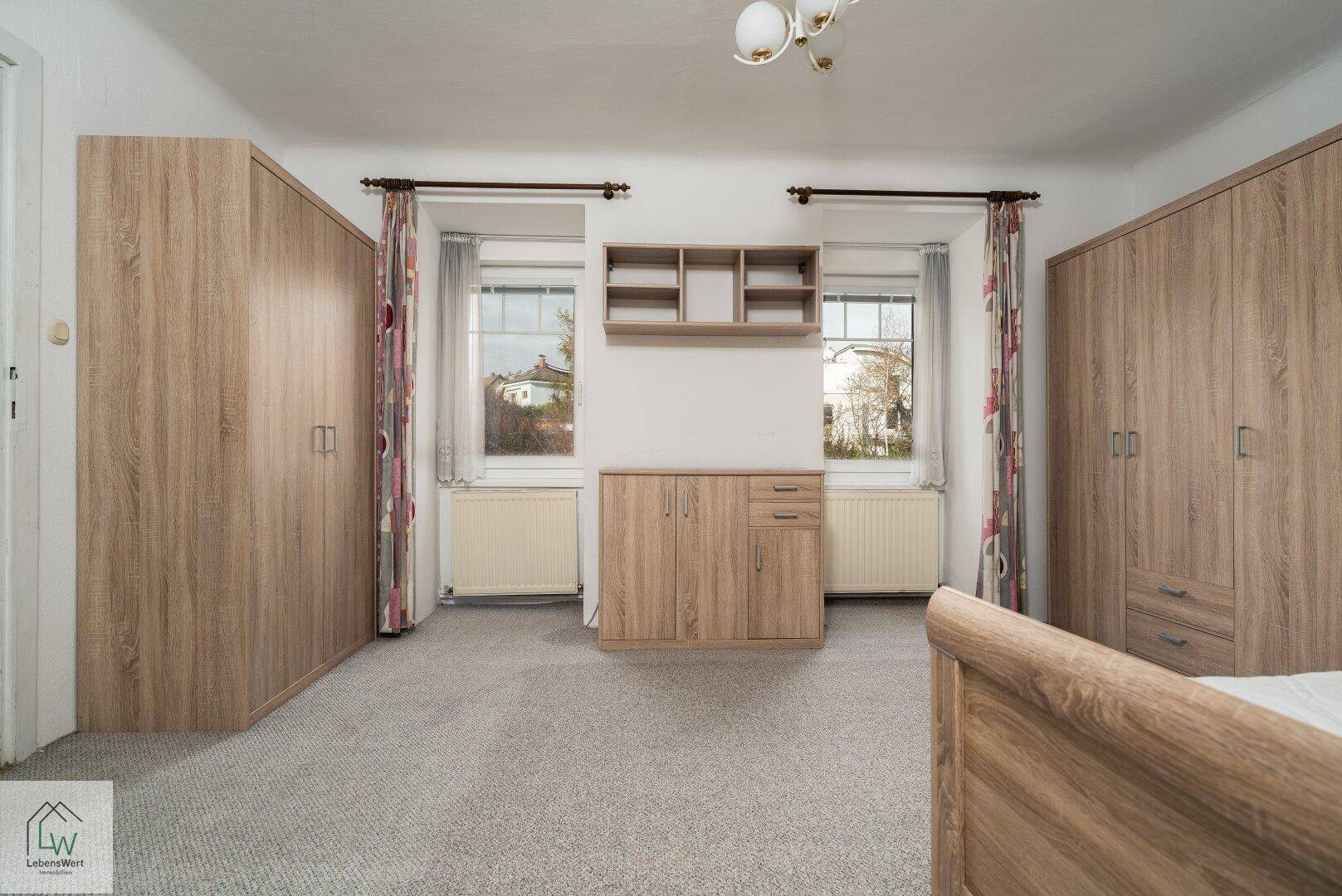Schlafzimmer mit Teppichboden, zwei Fenstern und Holzmöbeln, darunter zwei Kleiderschränke.