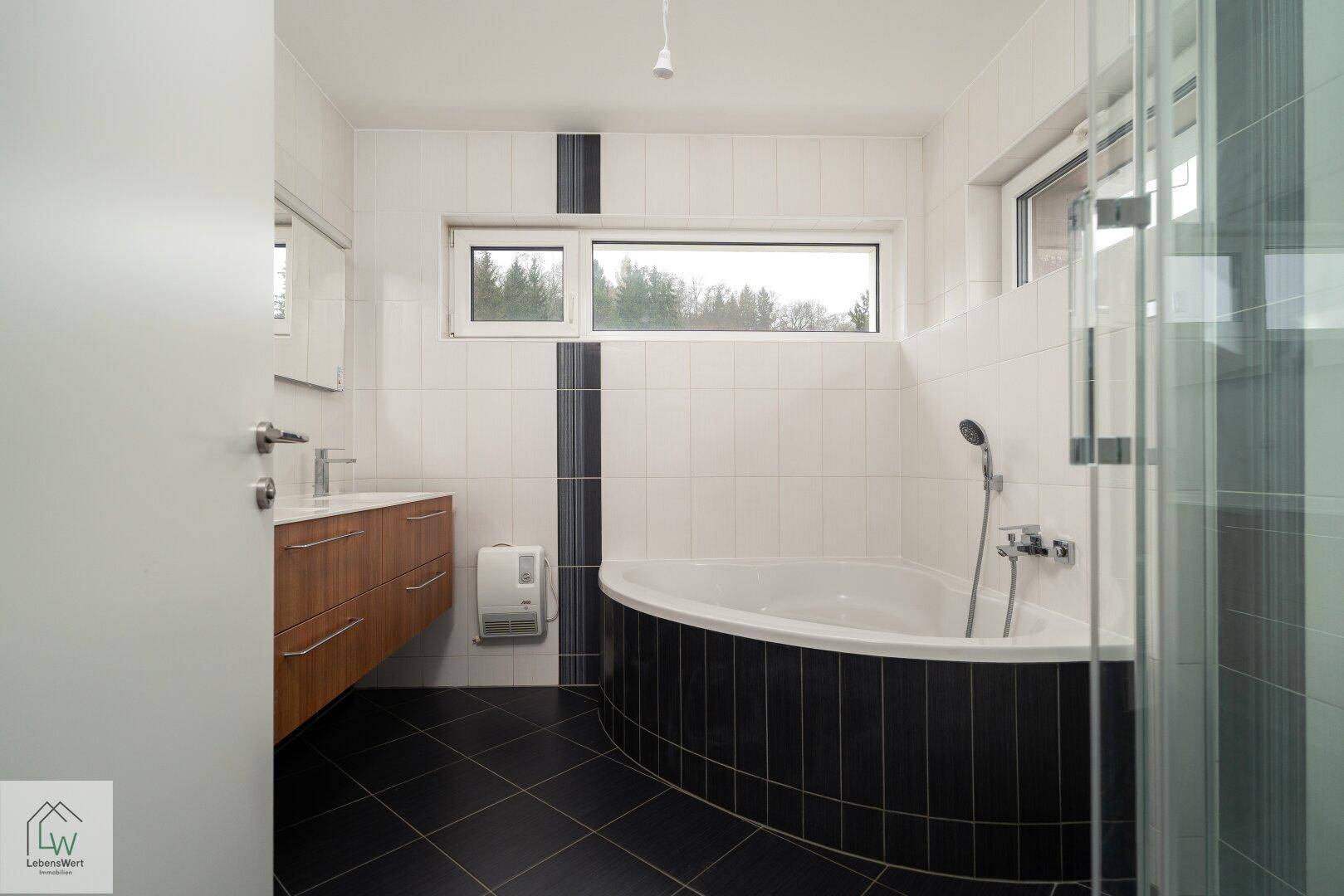 Modernes Badezimmer mit Eckbadewanne, Fenster und dunklen Bodenfliesen.