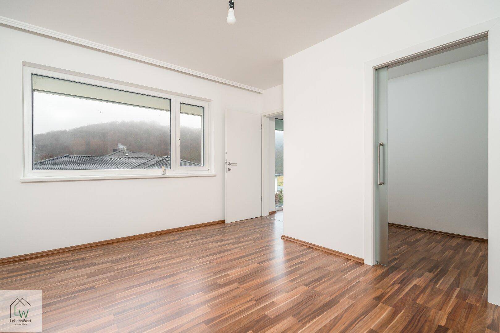 Helles Schlafzimmer mit Laminatboden, großem Fenster und Blick ins Grüne.