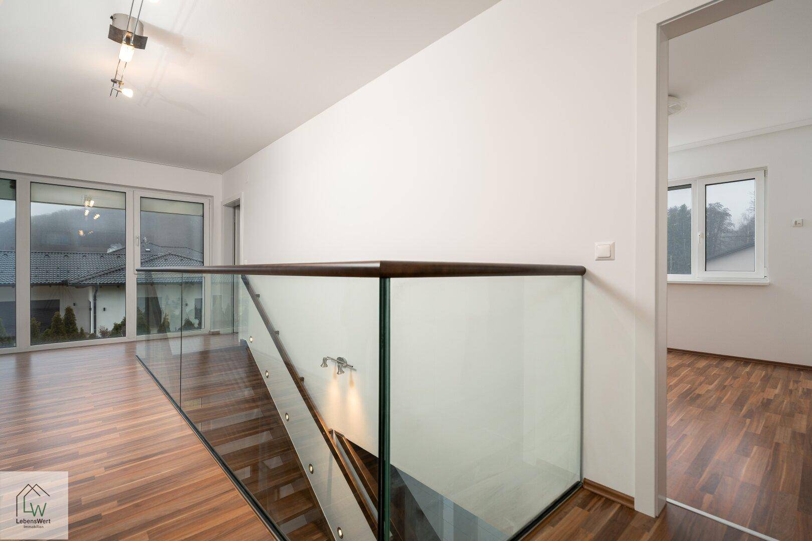 Heller Flur im Obergeschoss mit Glasgeländer, Laminatboden und großen Fenstern.