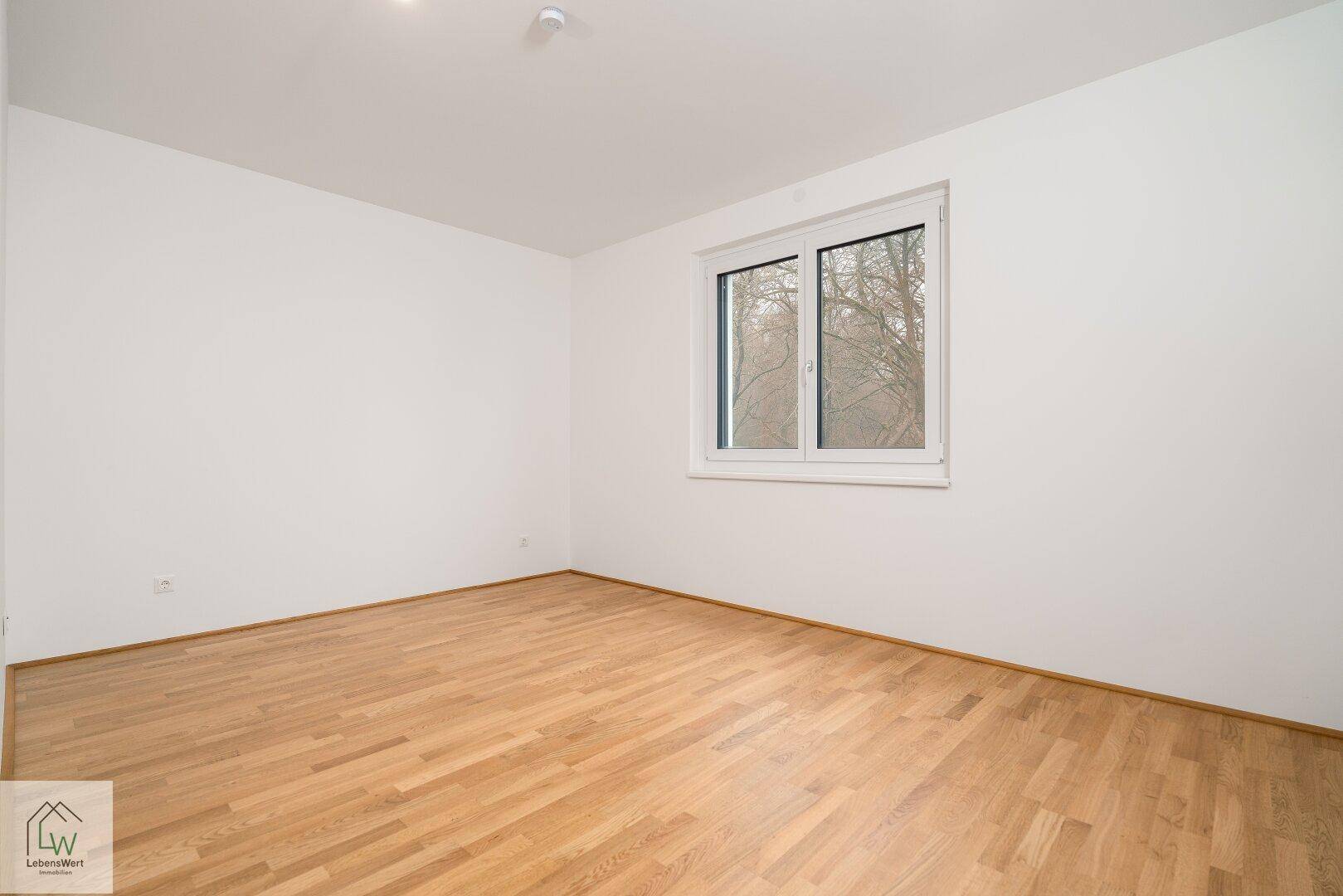 Leeres Zimmer mit hellem Parkettboden und Fenster mit Blick ins Grüne.