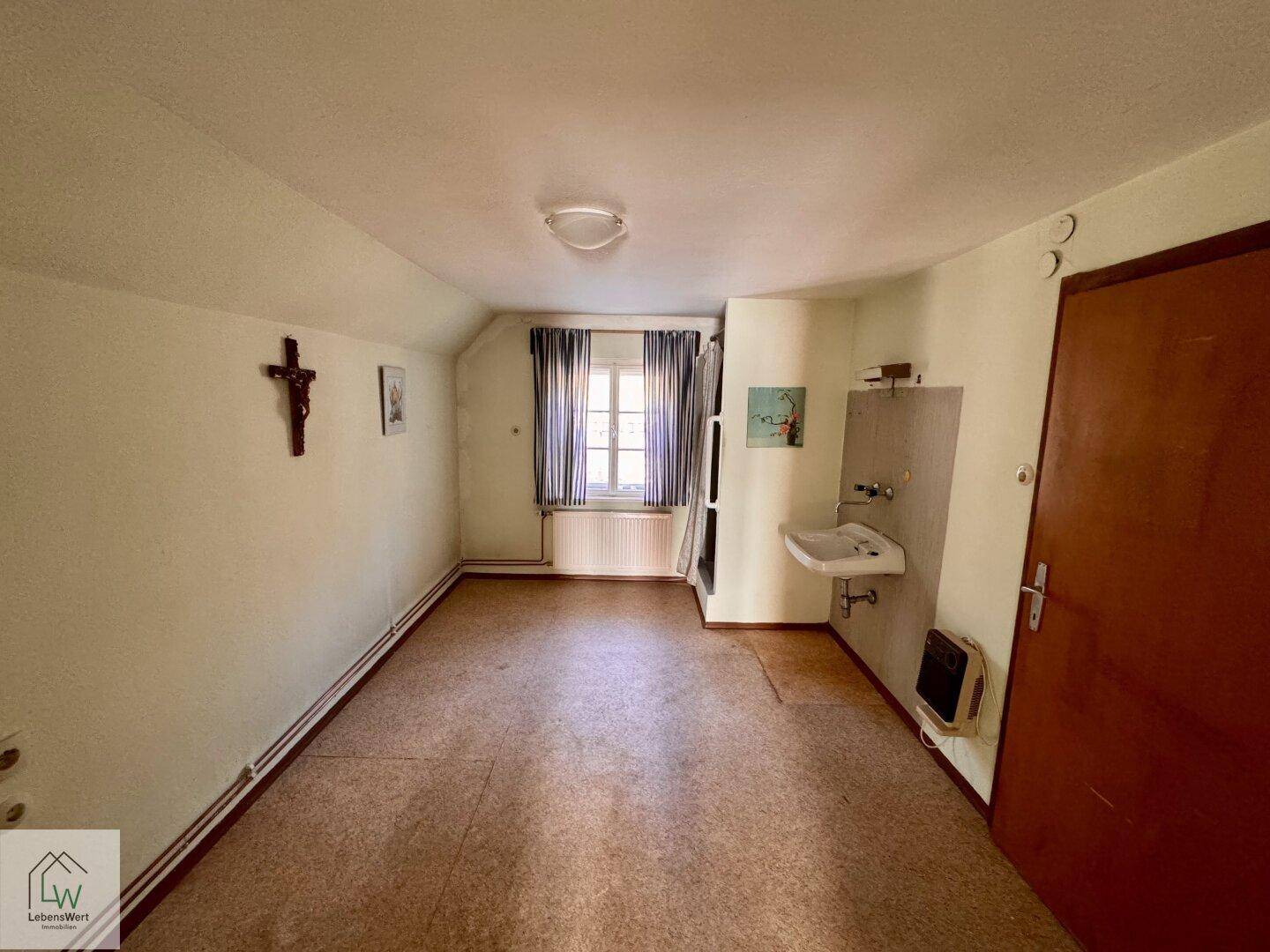 Einfacher Raum mit Waschbecken, Fenster und Teppichboden in einem älteren Zustand.