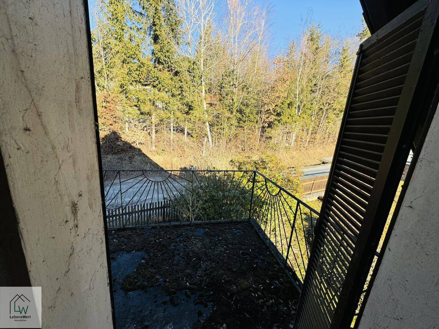 Alter Balkon mit schmiedeeisernem Geländer und Blick auf die bewaldete Umgebung.