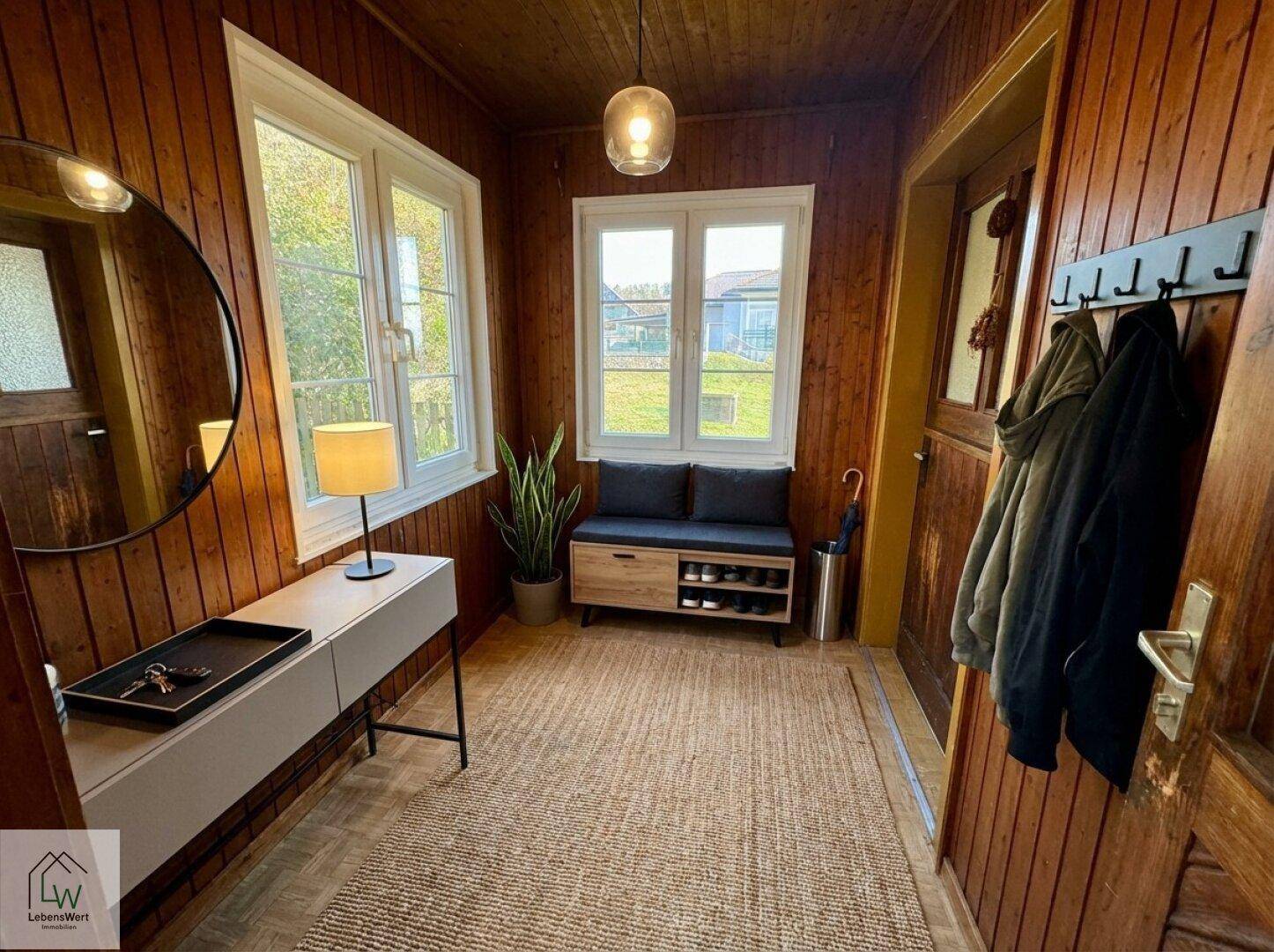 Gemütlicher Eingangsbereich mit Holzvertäfelung, Spiegel und Schuhbank, hell durch große Fenster.