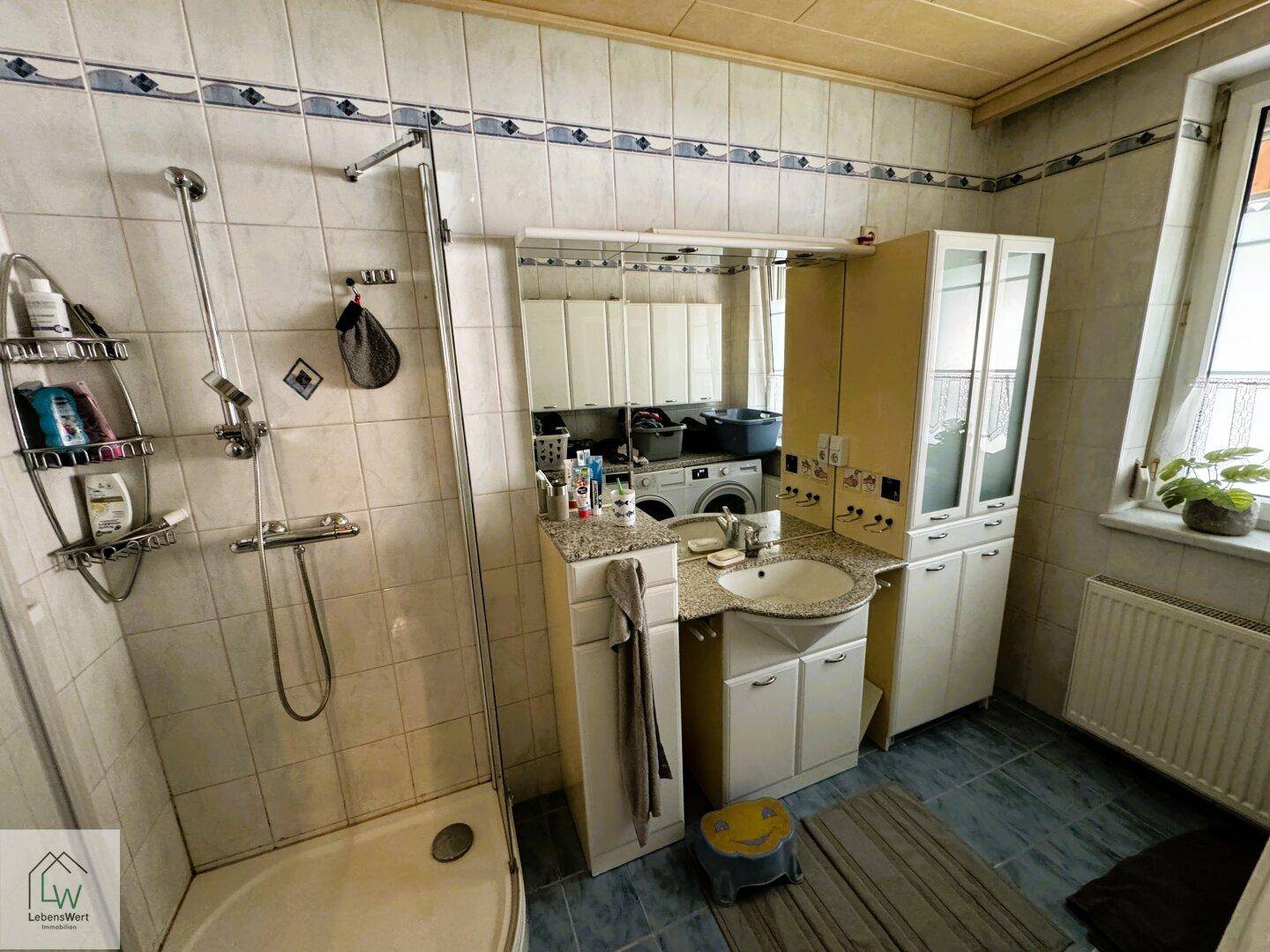 Badezimmer mit Duschkabine, Waschbecken, Waschmaschine und Fliesen an Wänden und Boden.