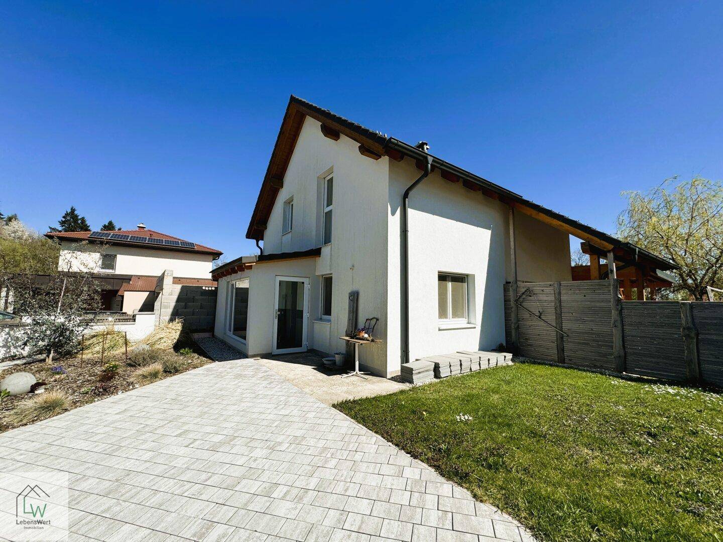 Seitenansicht des Hauses mit gepflasterter Auffahrt und kleinem Vorgarten unter blauem Himmel.