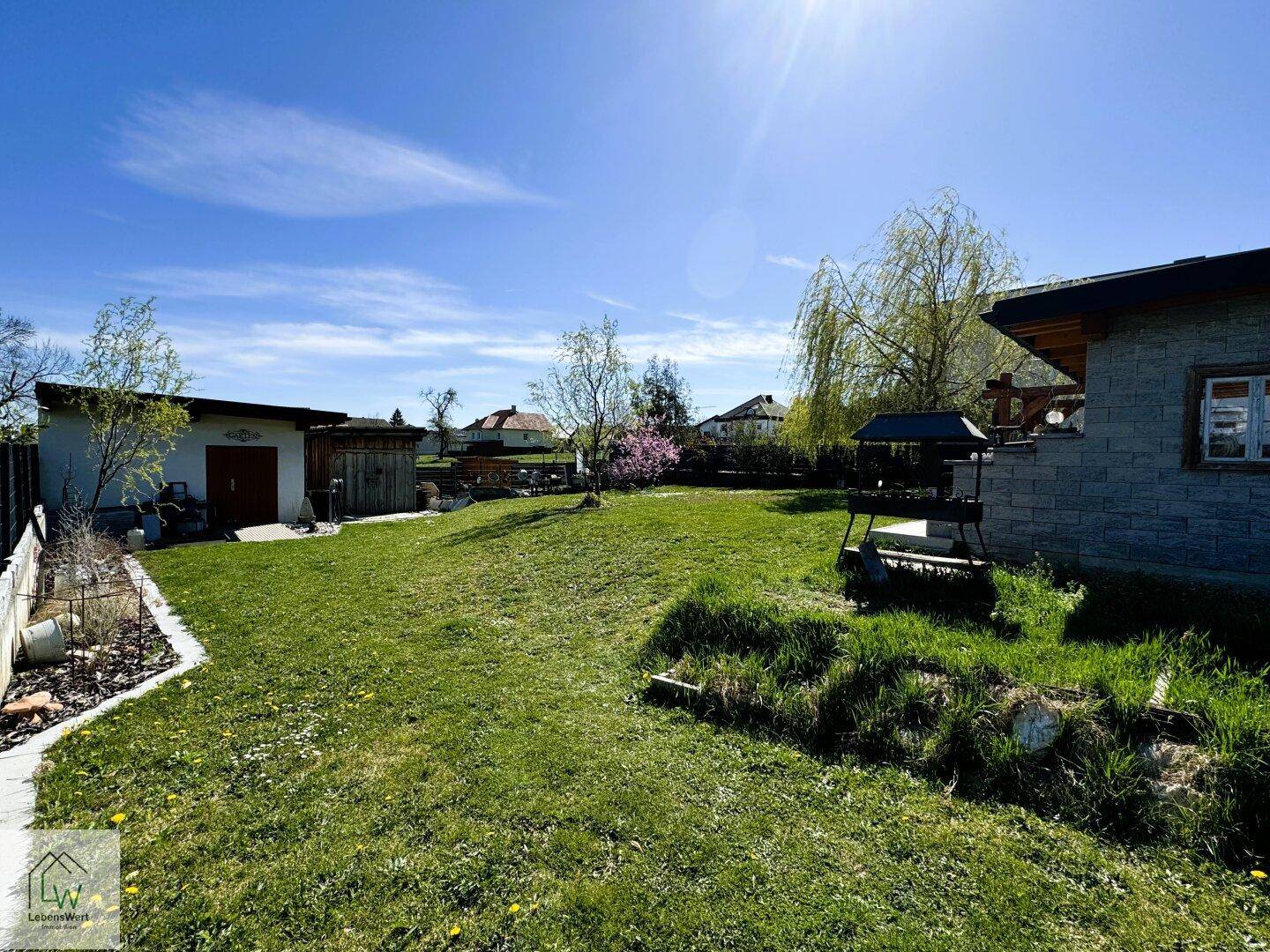 Großer, sonniger Garten mit Rasenfläche, Grillbereich und Gartenhaus im Hintergrund.
