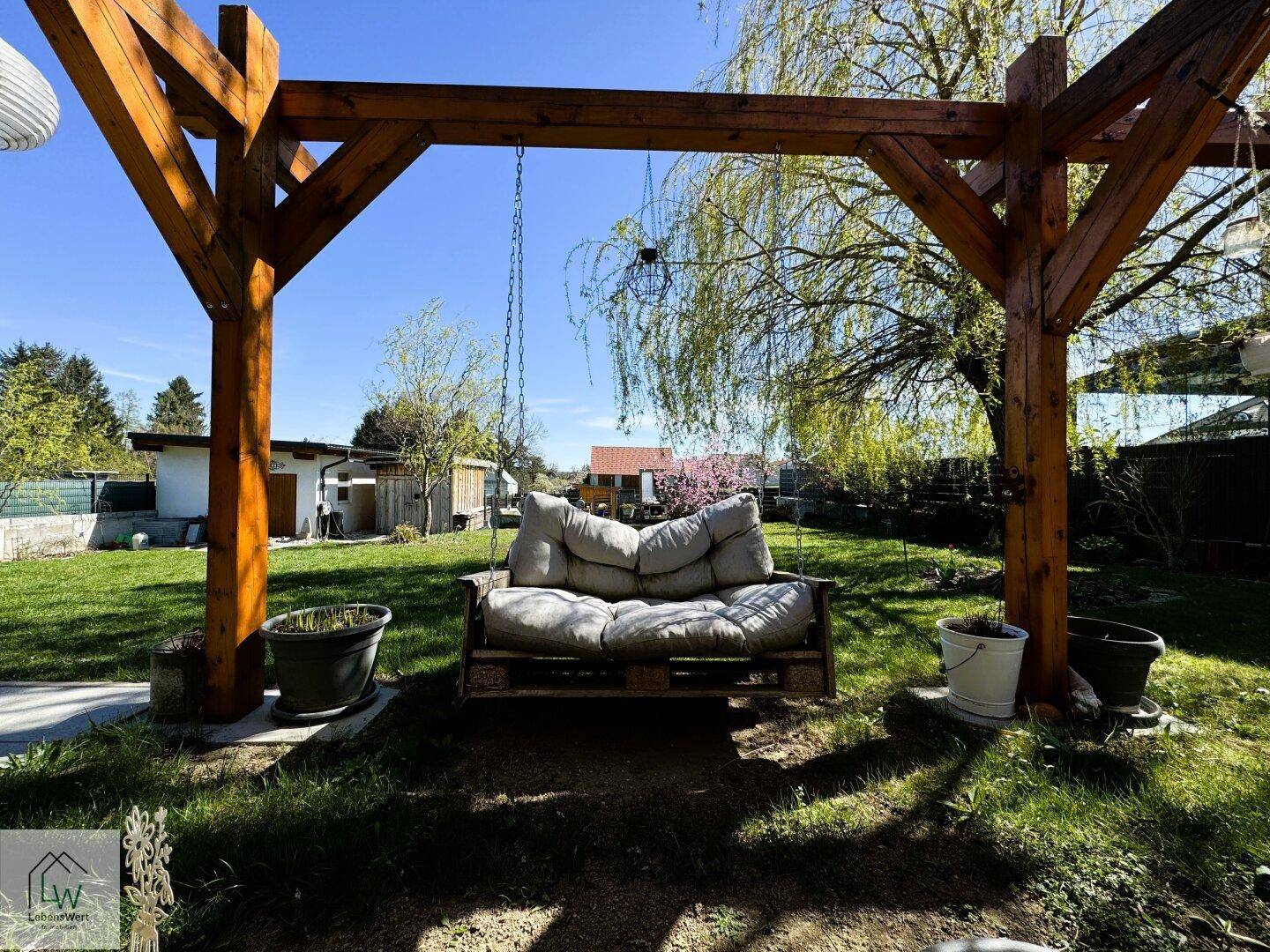 Schaukelbank unter einer Holzpergola im sonnigen Garten, umgeben von Grün.