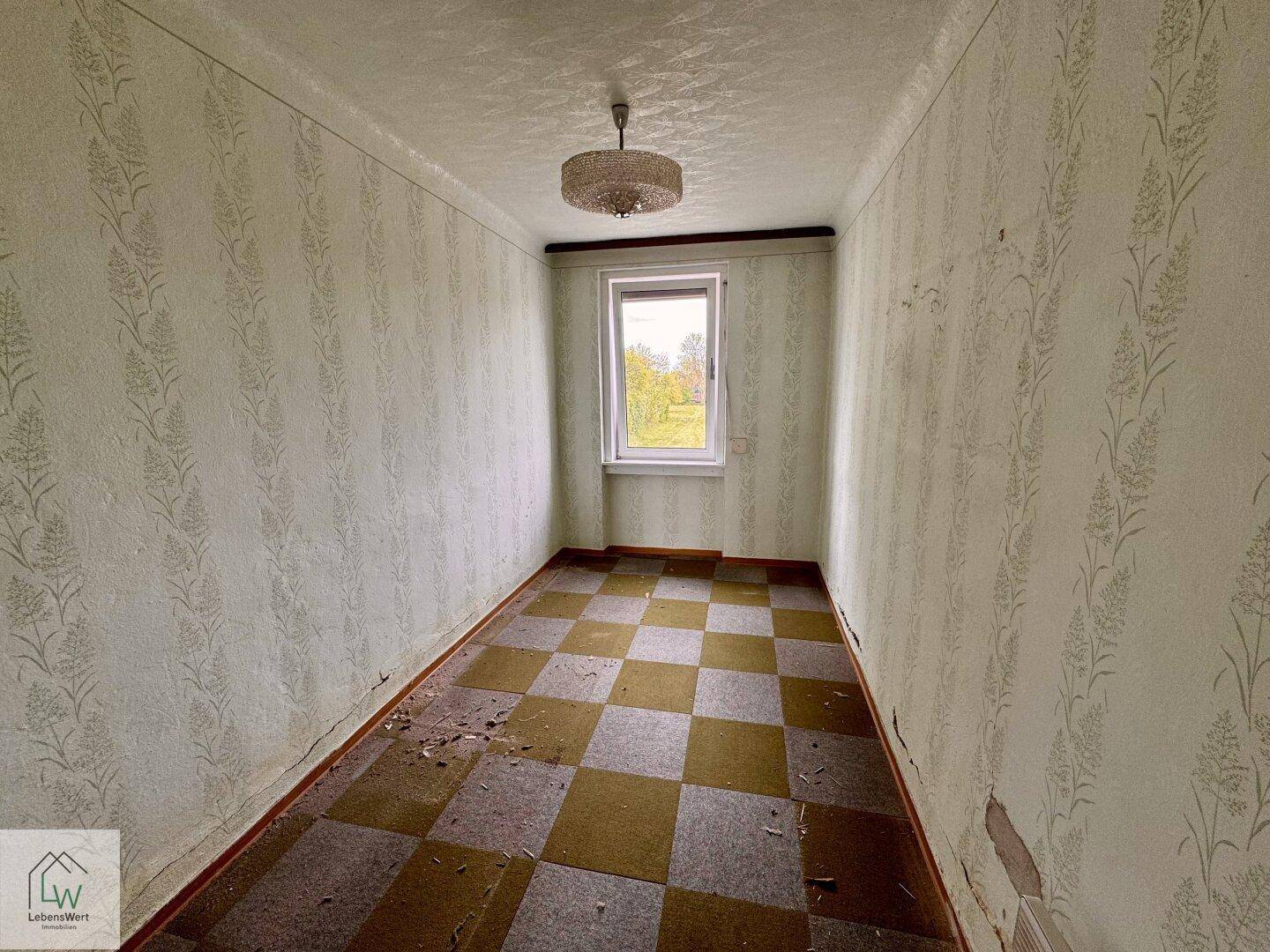 Kleines Schlafzimmer mit kariertem Teppichboden, Fenster und einfacher Deckenleuchte.