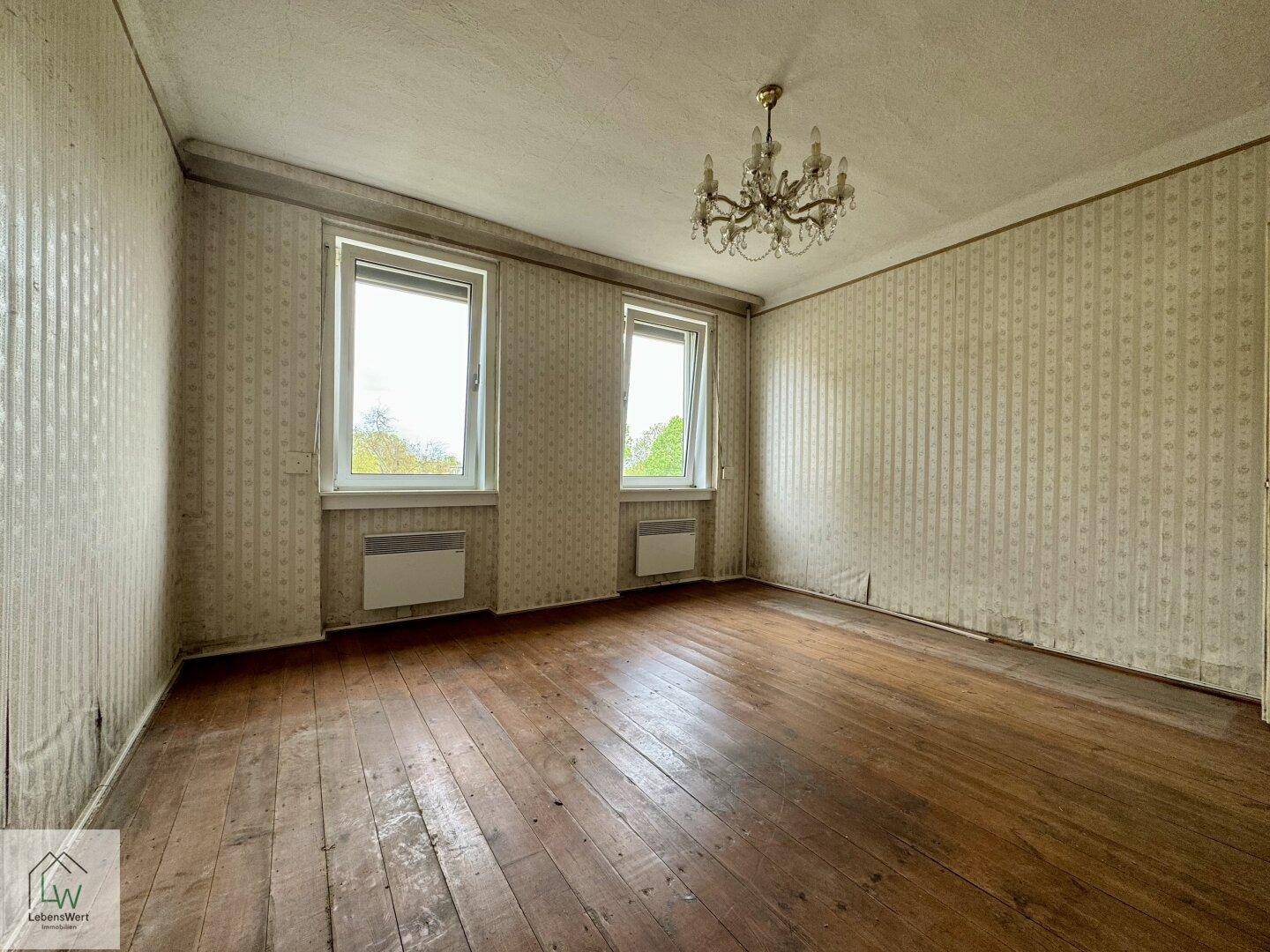 Geräumiges Wohnzimmer mit abgenutztem Holzdielenboden, Tapetenresten und Kronleuchter.