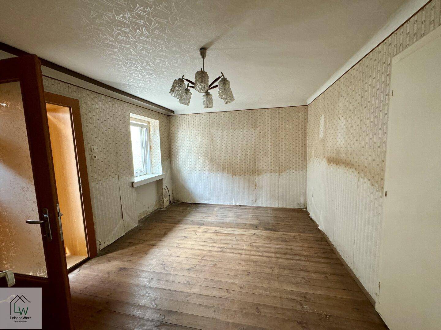 Helles Zimmer mit Holzdielenboden, beschädigter Tapete und einfacher Deckenleuchte.