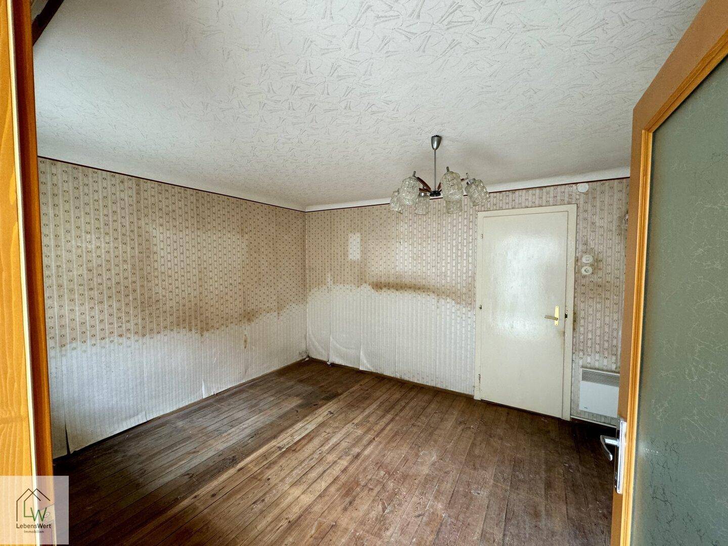 Geräumiger Raum mit Holzdielenboden, abgenutzter Tapete und einfacher Deckenleuchte.
