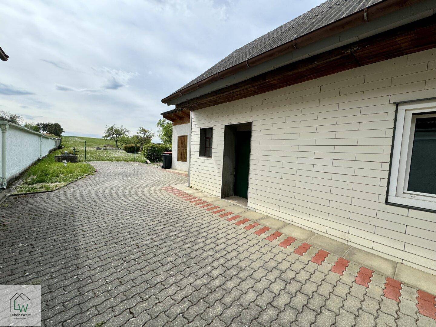 Gepflasterter Weg entlang der Hauswand, der zu einem grünen Bereich führt.