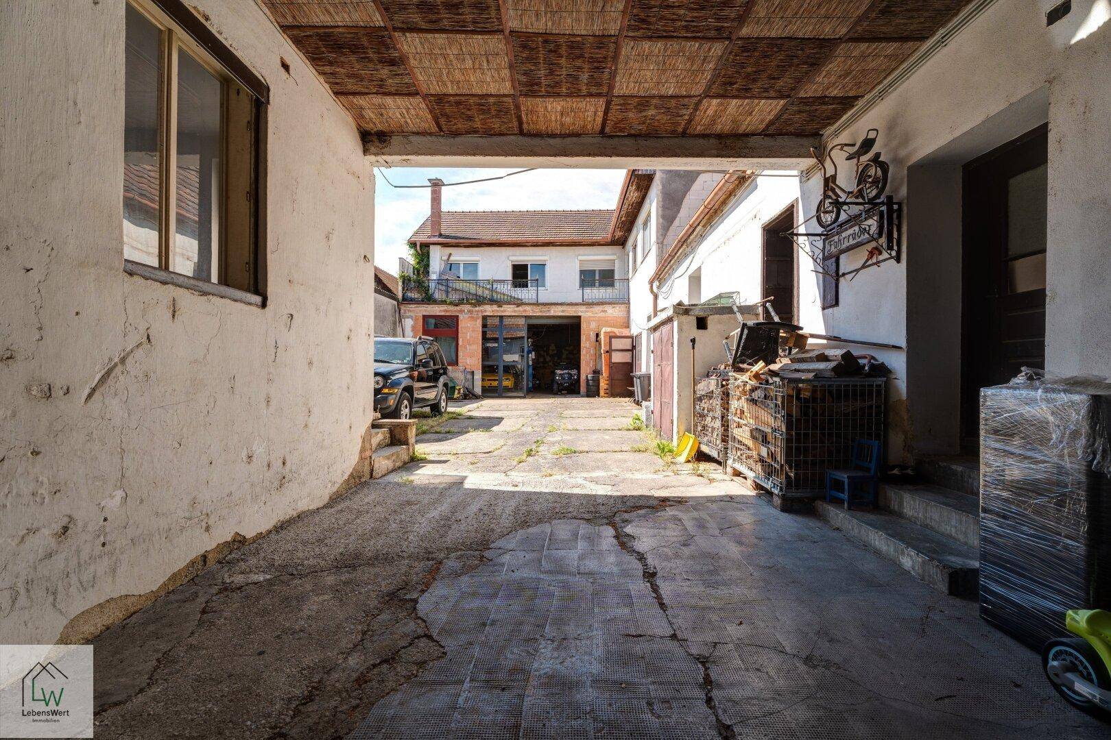 Überdachte Einfahrt zu einem Innenhof mit Blick auf weitere Gebäude und einen gepflasterten Bereich.