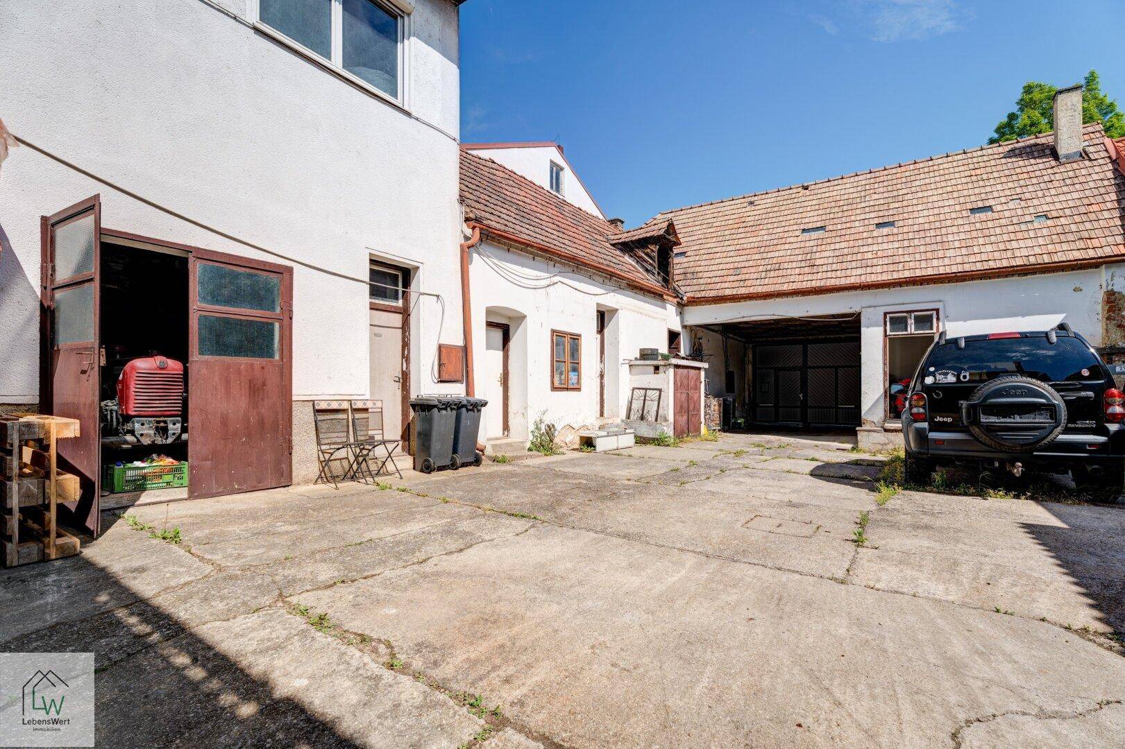 Innenhof mit offenem Zugang zu einer Werkstatt oder Garage und weiteren Gebäuden im Sonnenlicht.