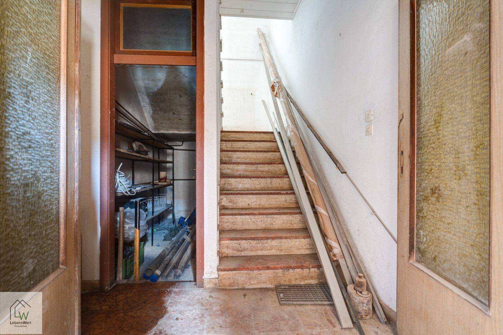 Flur mit Holztreppe und Zugang zu einem Abstellraum, der Renovierungsbedarf zeigt.