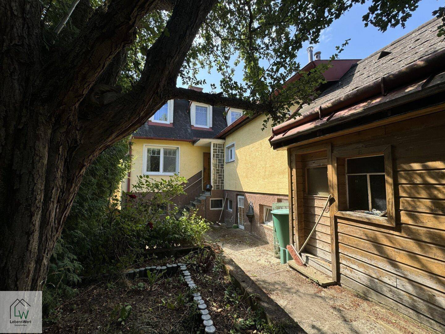Seitenansicht des Hauses mit einem rustikalen Holzschuppen und altem Baumbestand im Garten.