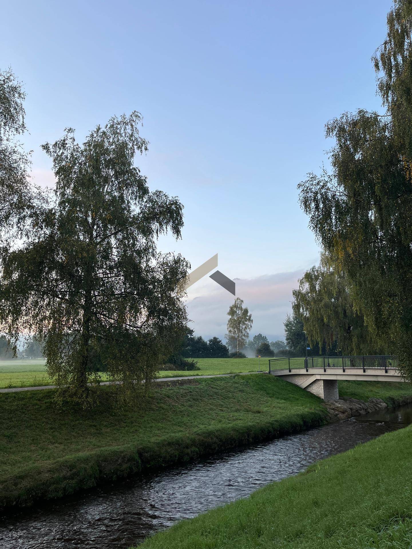 Ruhige Flusslandschaft mit Brücke, Bäumen und grünen Ufern in nebliger Morgenstimmung.
