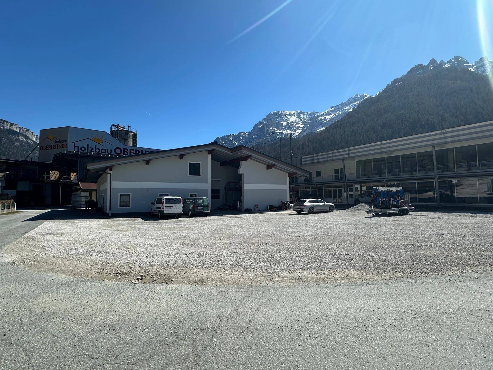 Gewerbegebäude mit großem Vorplatz und schneebedeckten Bergen im Hintergrund unter strahlendem Sonnenschein.