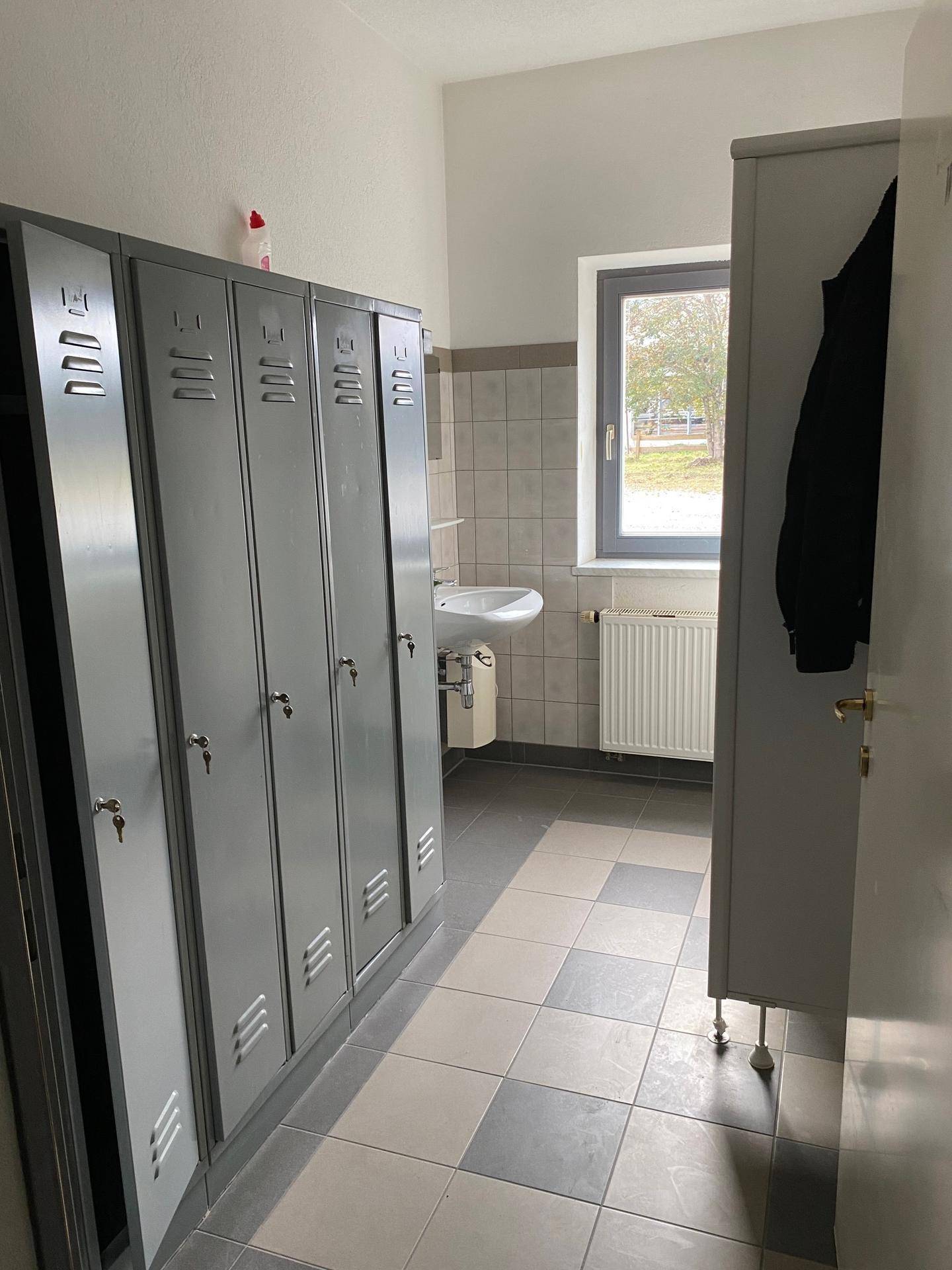 Umkleideraum mit Spinden, Waschbecken und einem Fenster, das Tageslicht hereinlässt und belüftet.