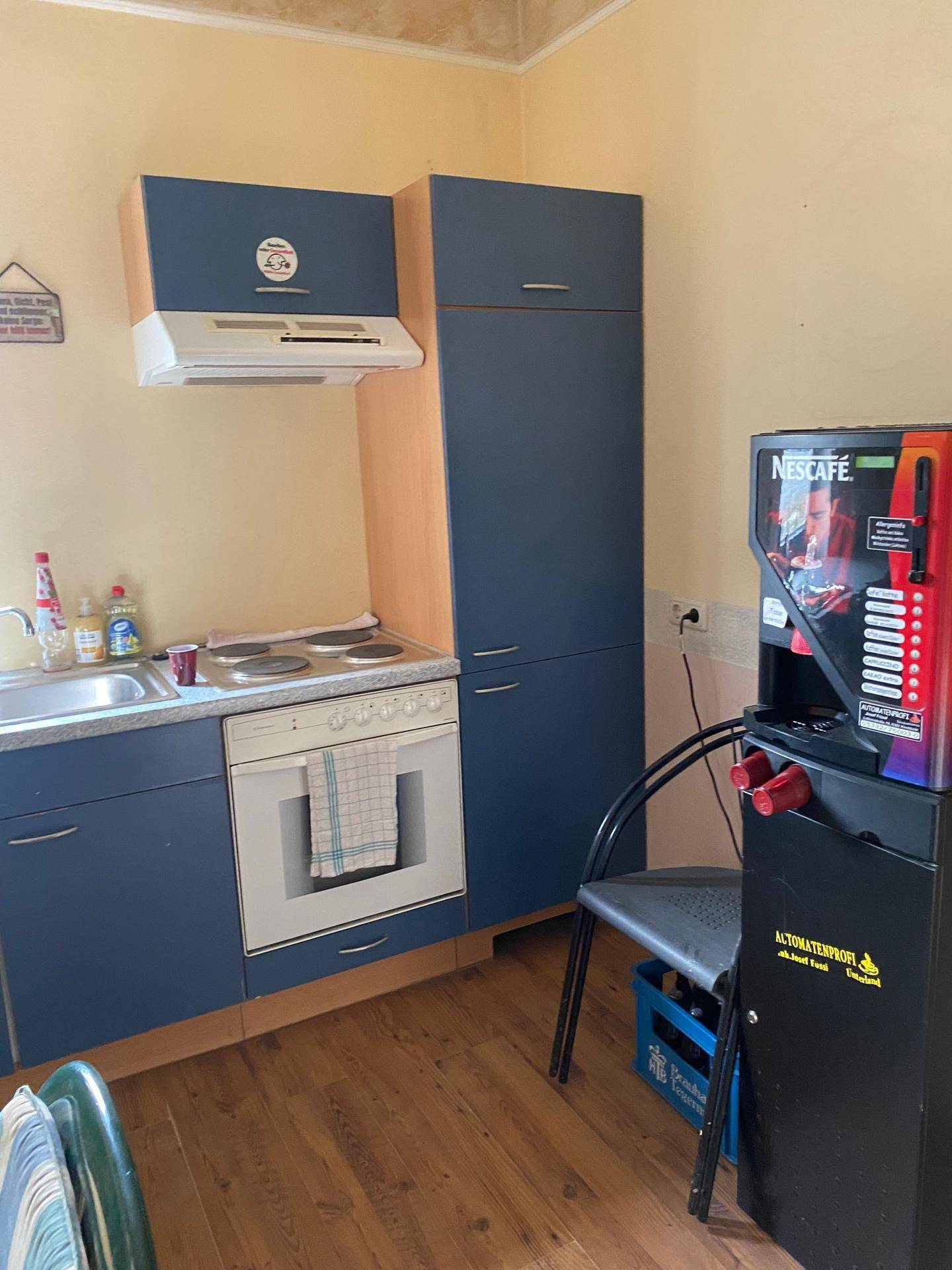 Kleine Küche mit blauen Schränken, Herd und Backofen, sowie einem praktischen Kaffeeautomaten.