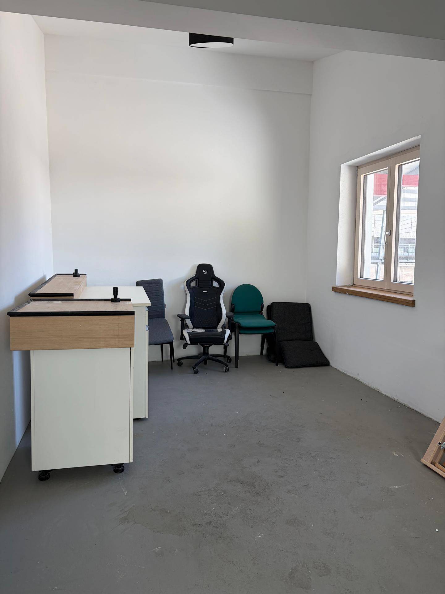 Einfacher Büroraum mit weißen Wänden, Betonboden und einem Fenster, das für Helligkeit sorgt.