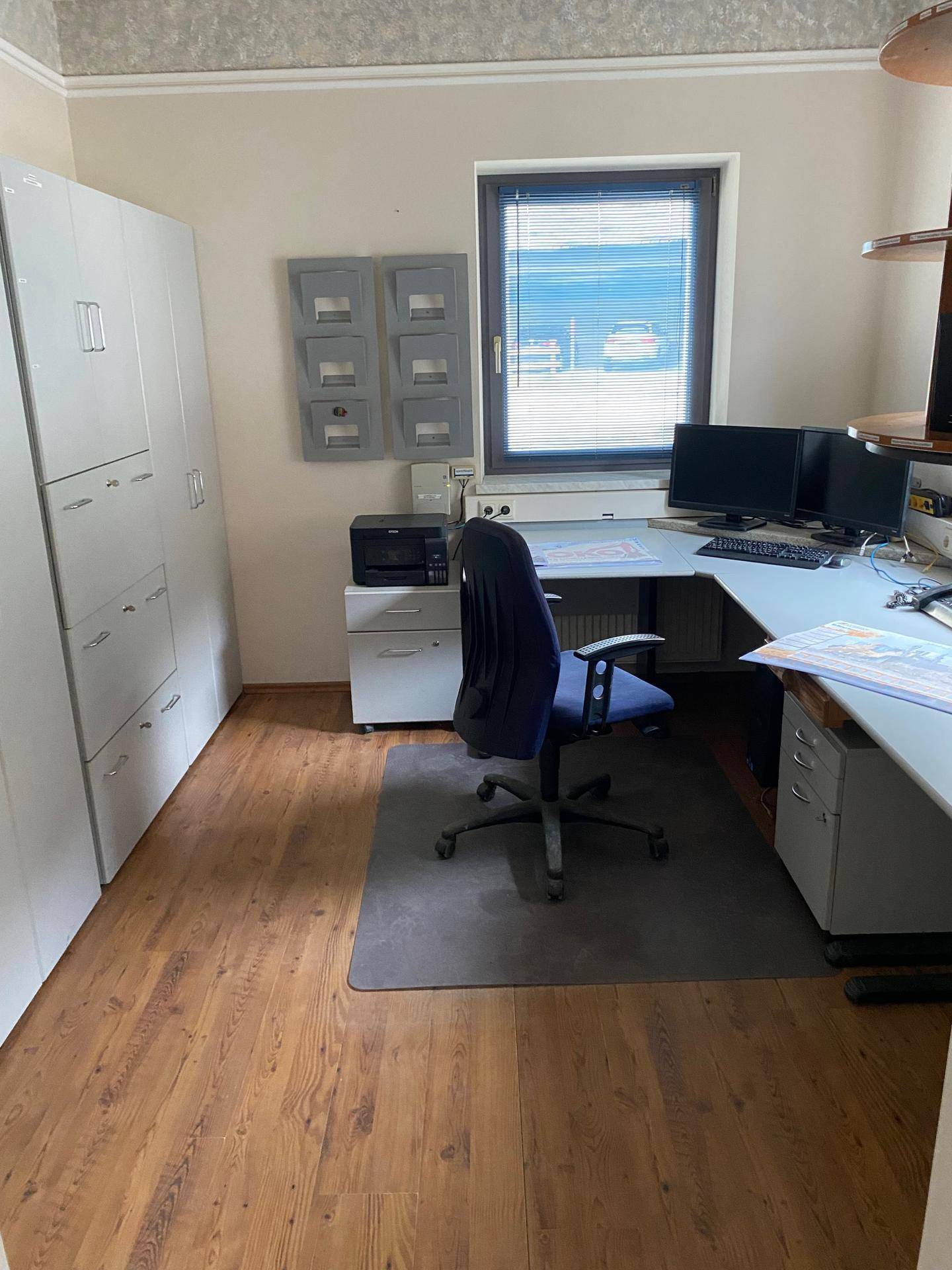 Helles Büro mit Holzfußboden, Schreibtisch und Aktenschränken, gut beleuchtet durch ein Fenster.
