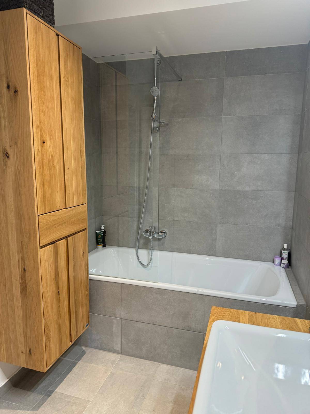 Badezimmer mit Badewanne, grauen Fliesen und einem hohen Holzschrank.