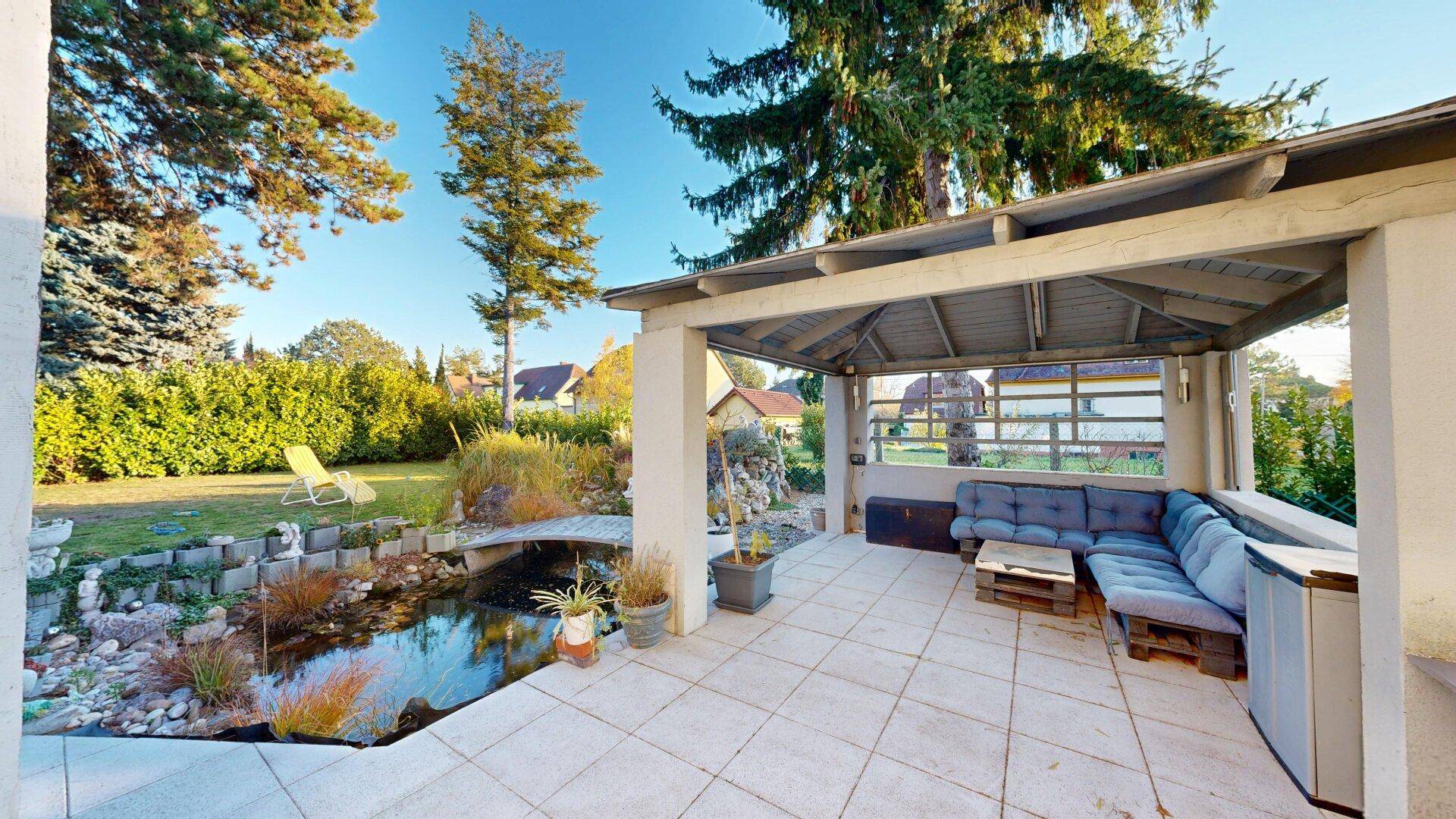 Überdachte Terrasse mit Fliesenboden, bequemen Sitzmöbeln und Blick auf den Teich und Garten.