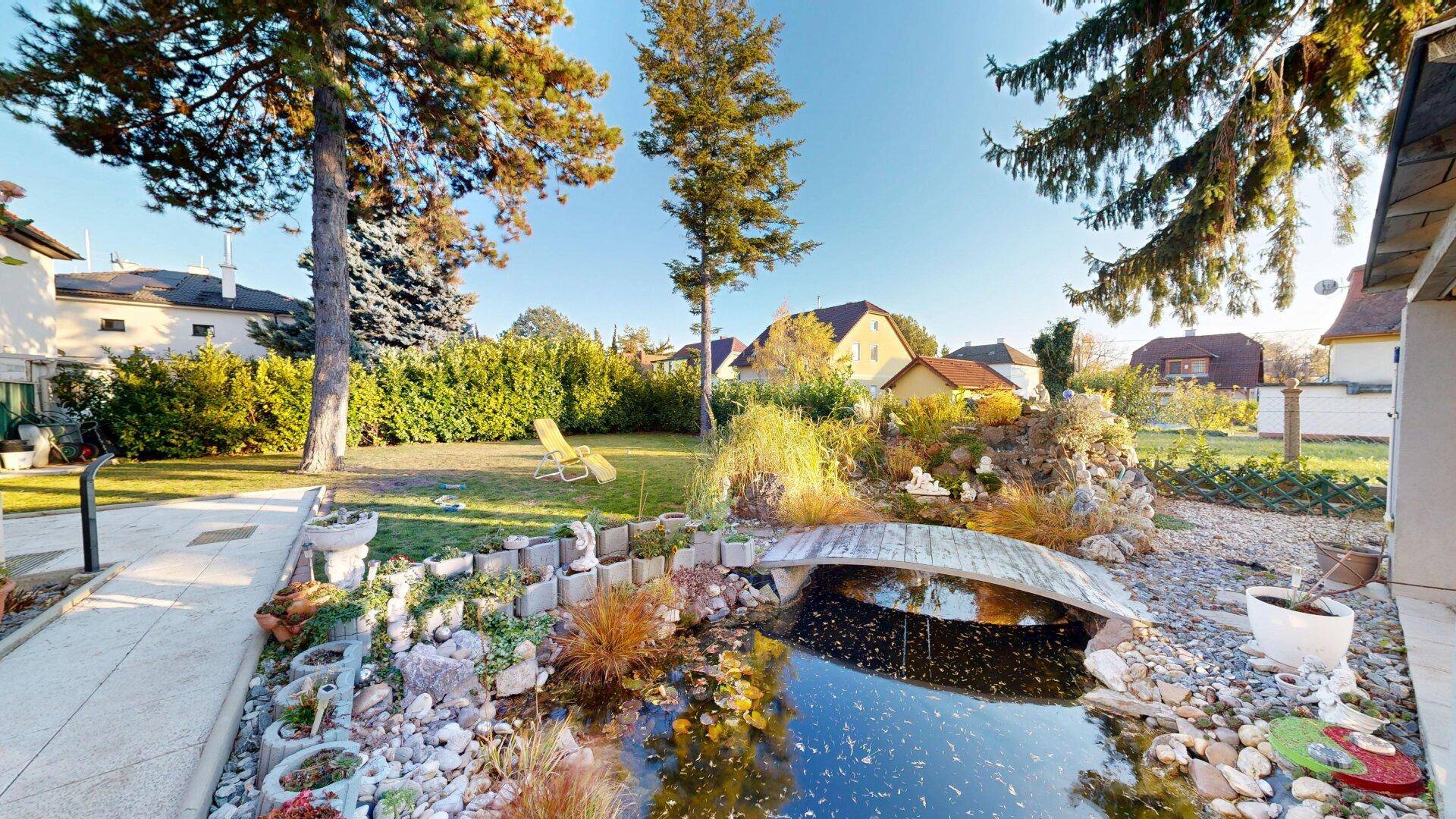 Idyllischer Gartenbereich mit einem Teich, einer kleinen Brücke und üppiger Bepflanzung.