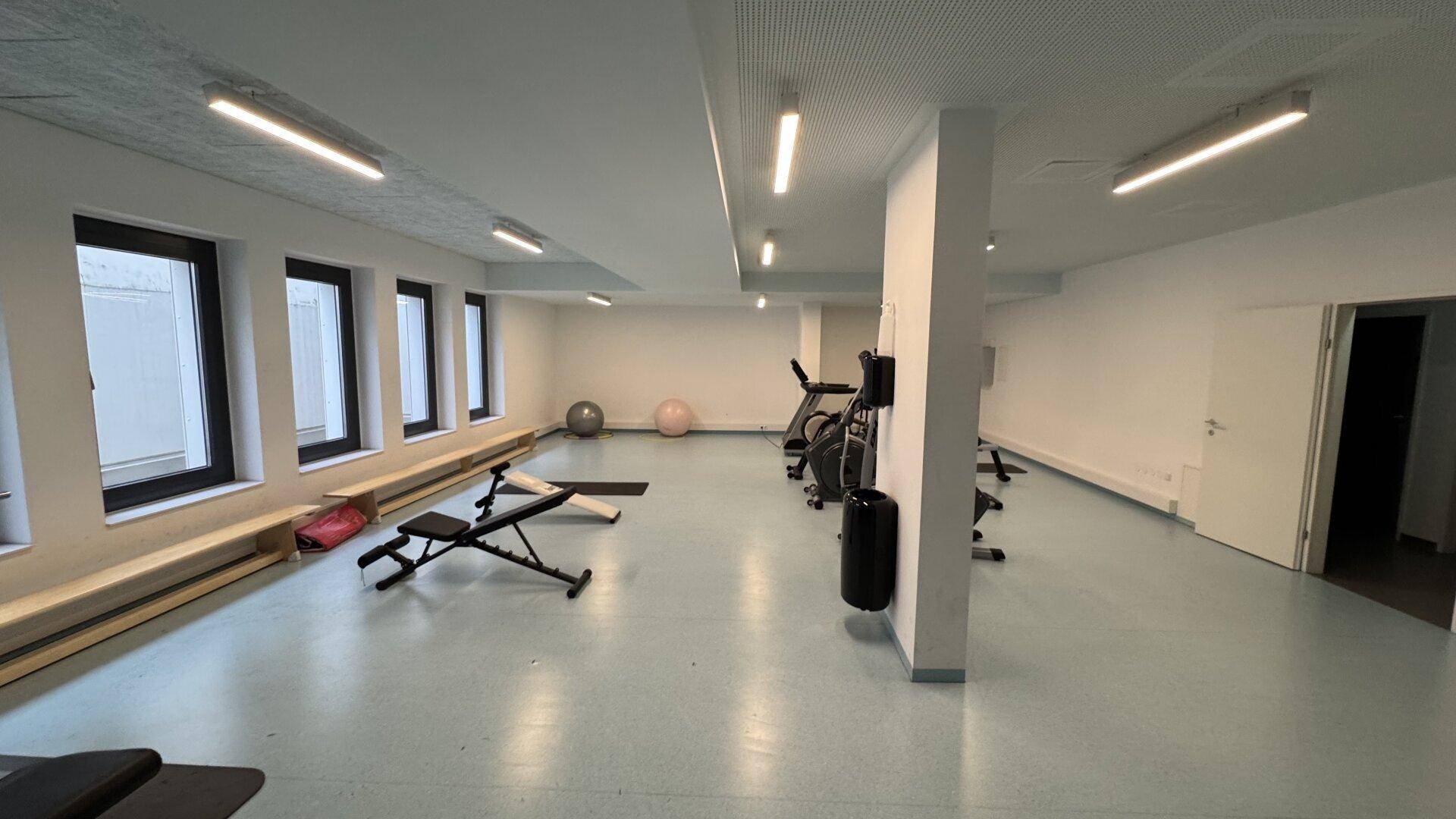 Gemeinschaftlicher Fitnessraum mit verschiedenen Trainingsgeräten und großen Fenstern.