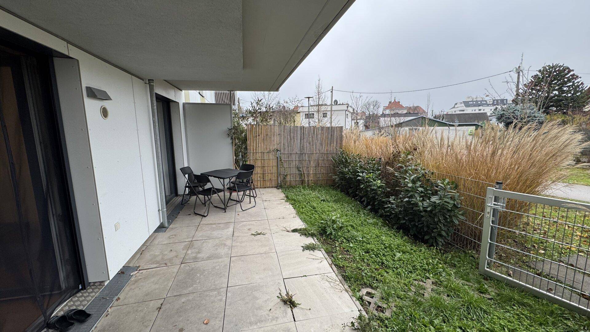 Geflieste Terrasse mit Gartentisch und Stühlen, umgeben von Pflanzen und einem Zaun.