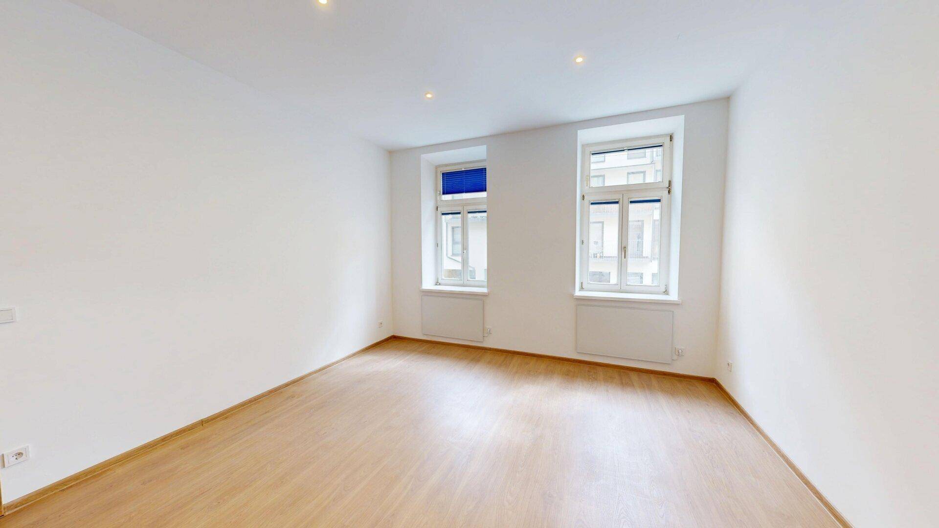 Geräumiges, helles Zimmer mit zwei großen Fenstern und Holzfußboden, ideal für Wohnzwecke.