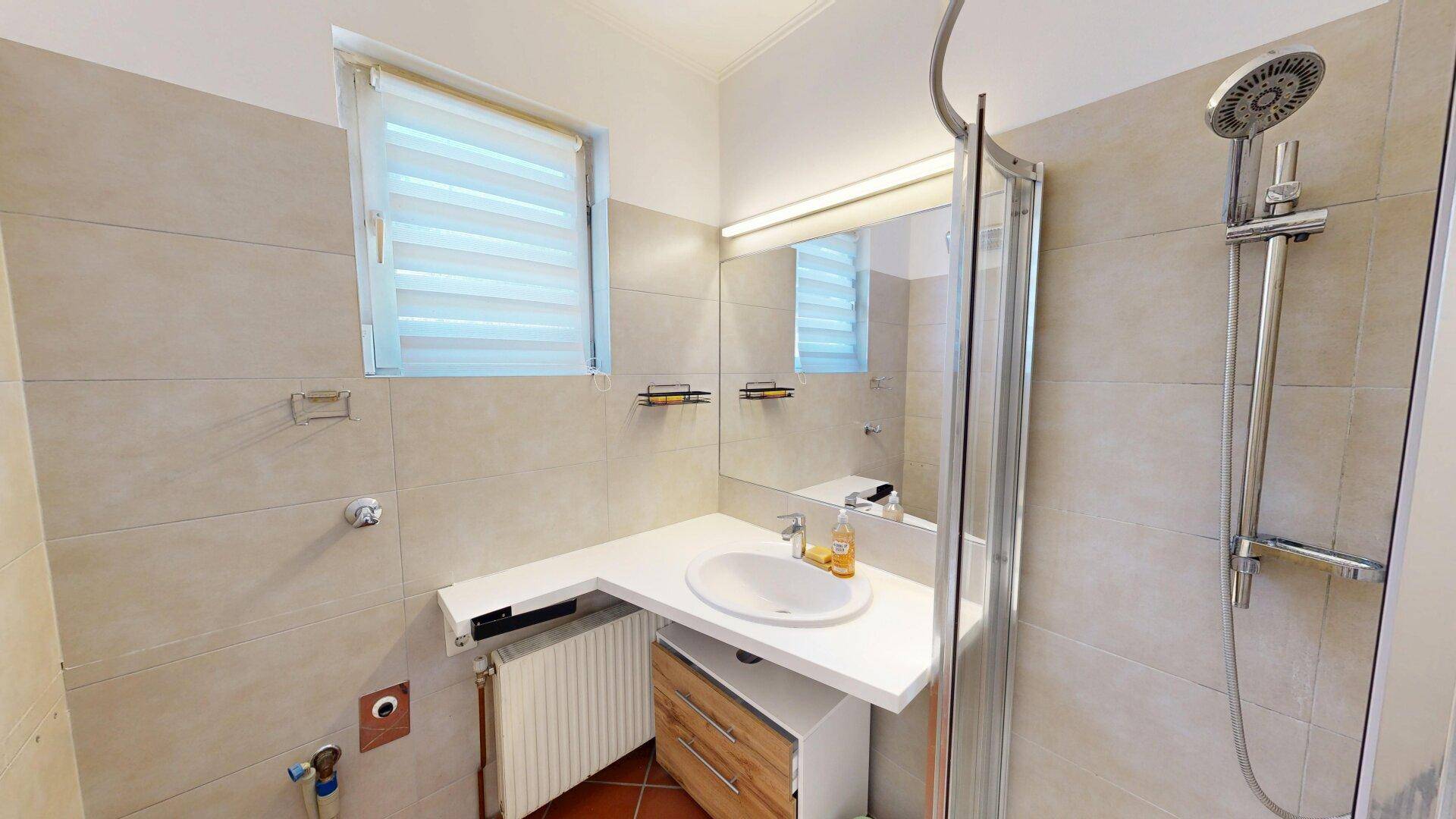 Badezimmer mit Dusche, Waschbecken, Fenster und beigefarbenen Fliesen.