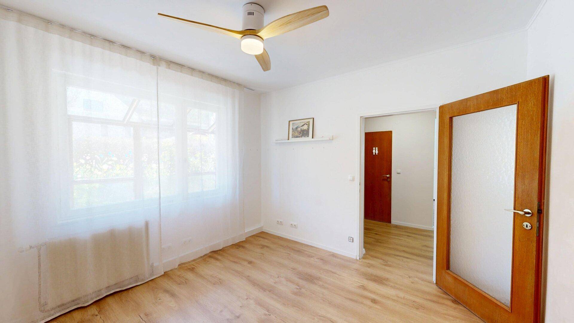 Schlafzimmer mit hellem Laminatboden, großem Fenster und Deckenventilator für angenehmes Klima.