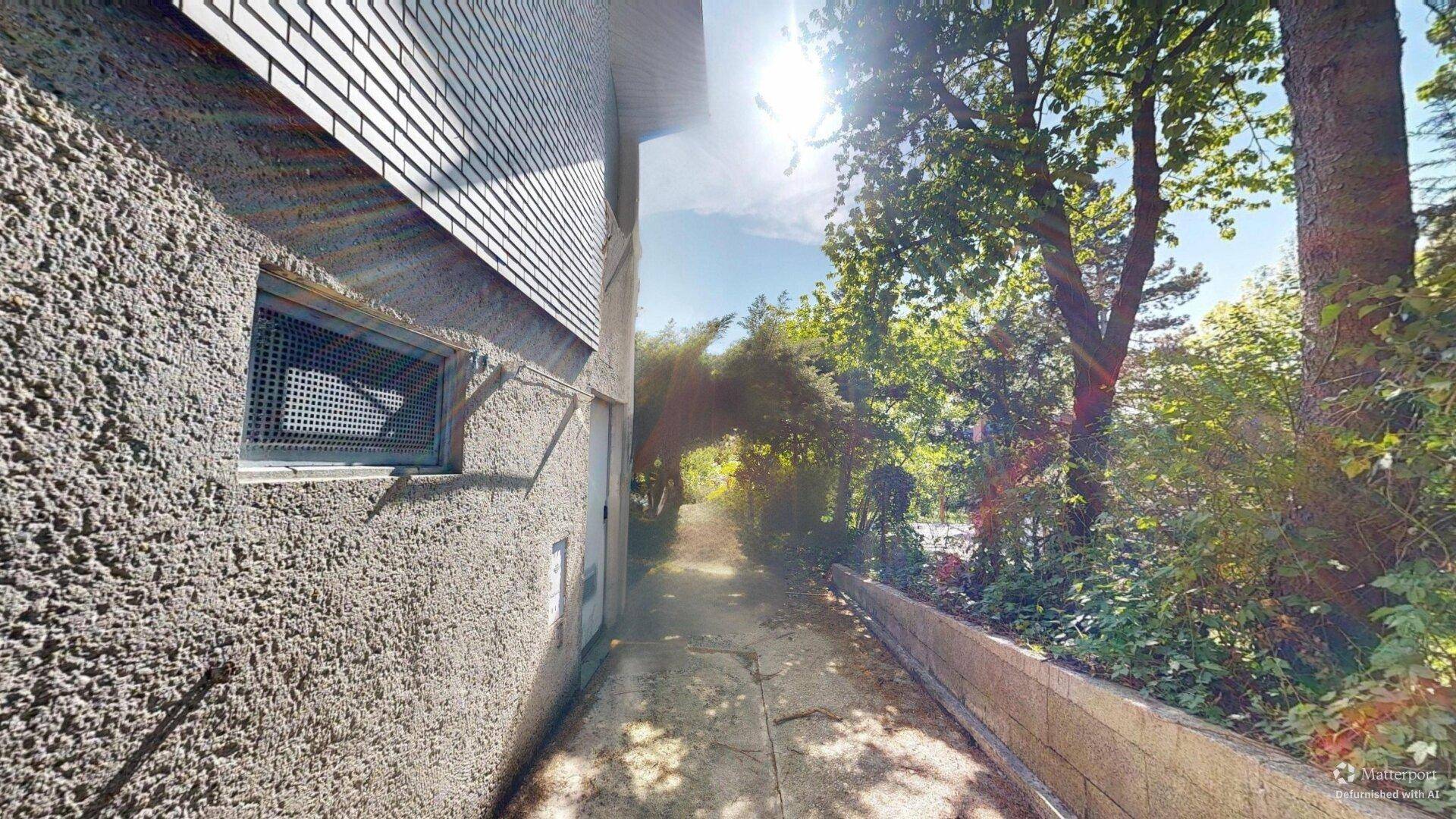 Schmaler Weg entlang des Hauses mit viel Grün und Bäumen im Sonnenlicht.