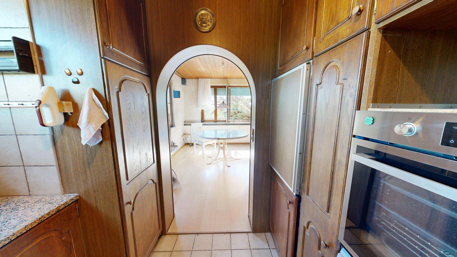 Küche mit Holzschränken und Durchgang zum Wohnbereich, mit integriertem Backofen.