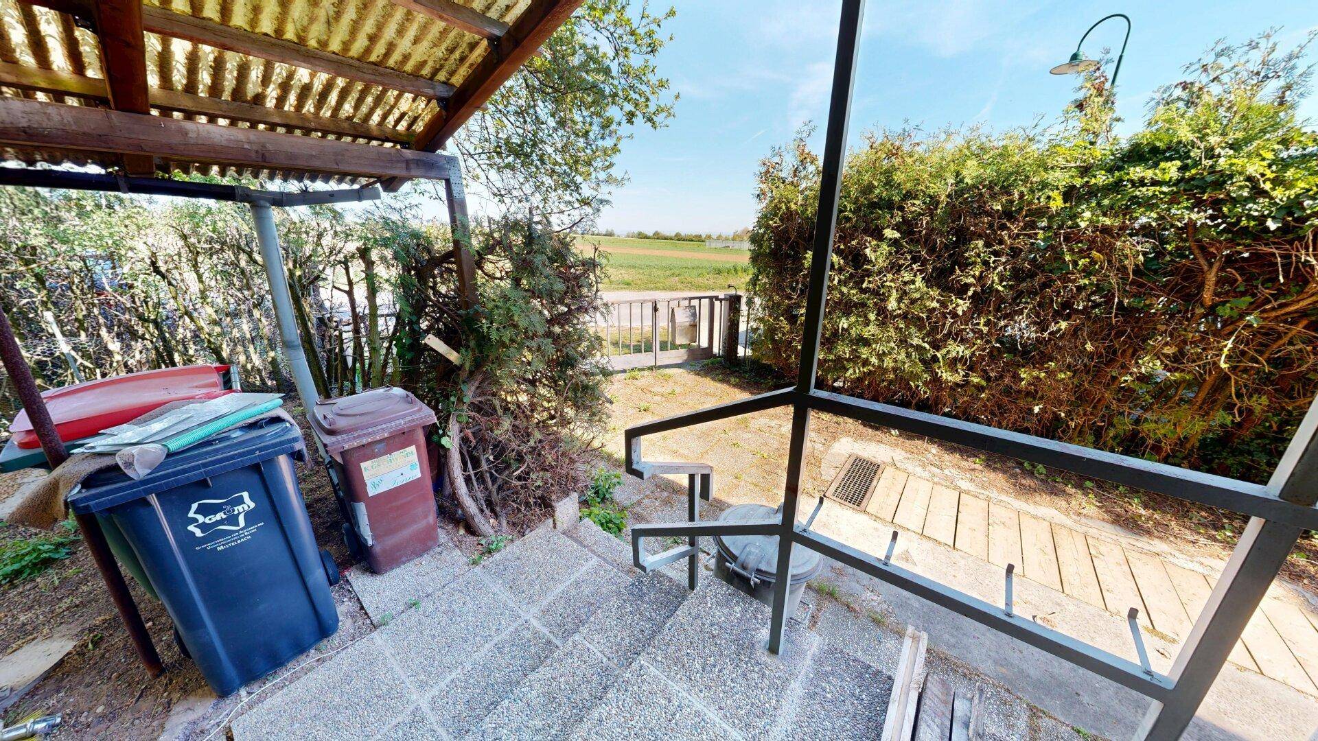 Eingangsbereich mit Treppe, Mülltonnen und Blick auf die umliegende Landschaft.