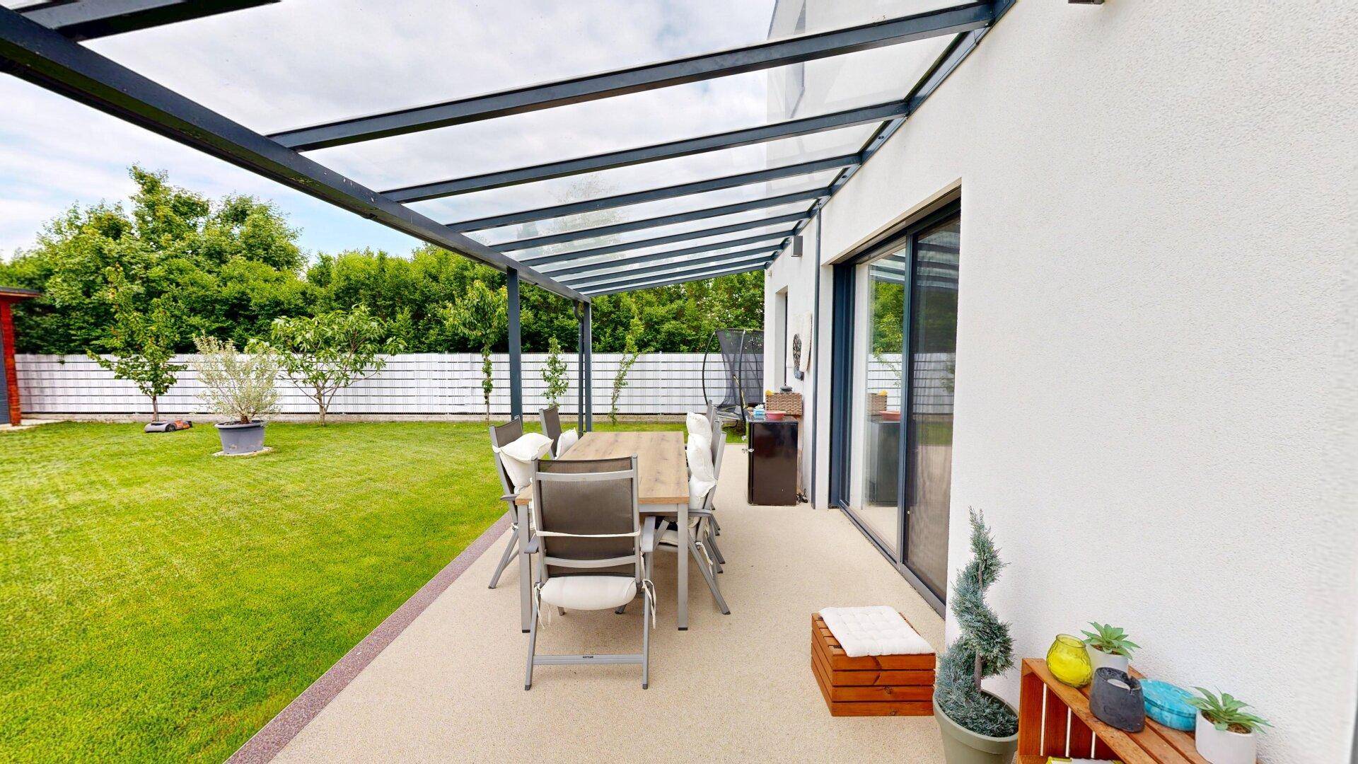 Überdachte Terrasse mit Esstisch und Stühlen, umgeben von einem grünen Garten.
