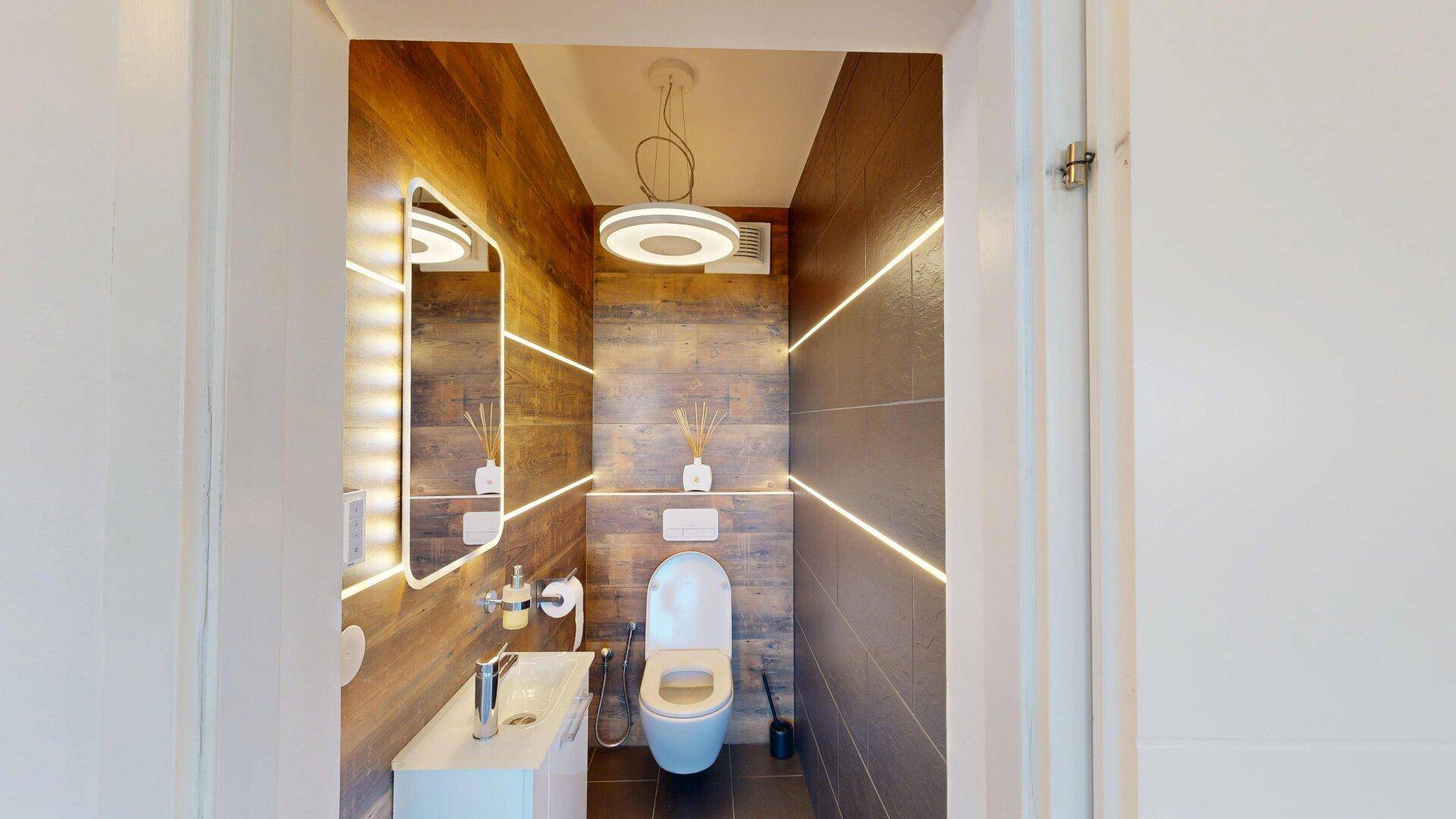 Modernes Gäste-WC mit dunklen Fliesen, Wand-WC und beleuchtetem Spiegel.