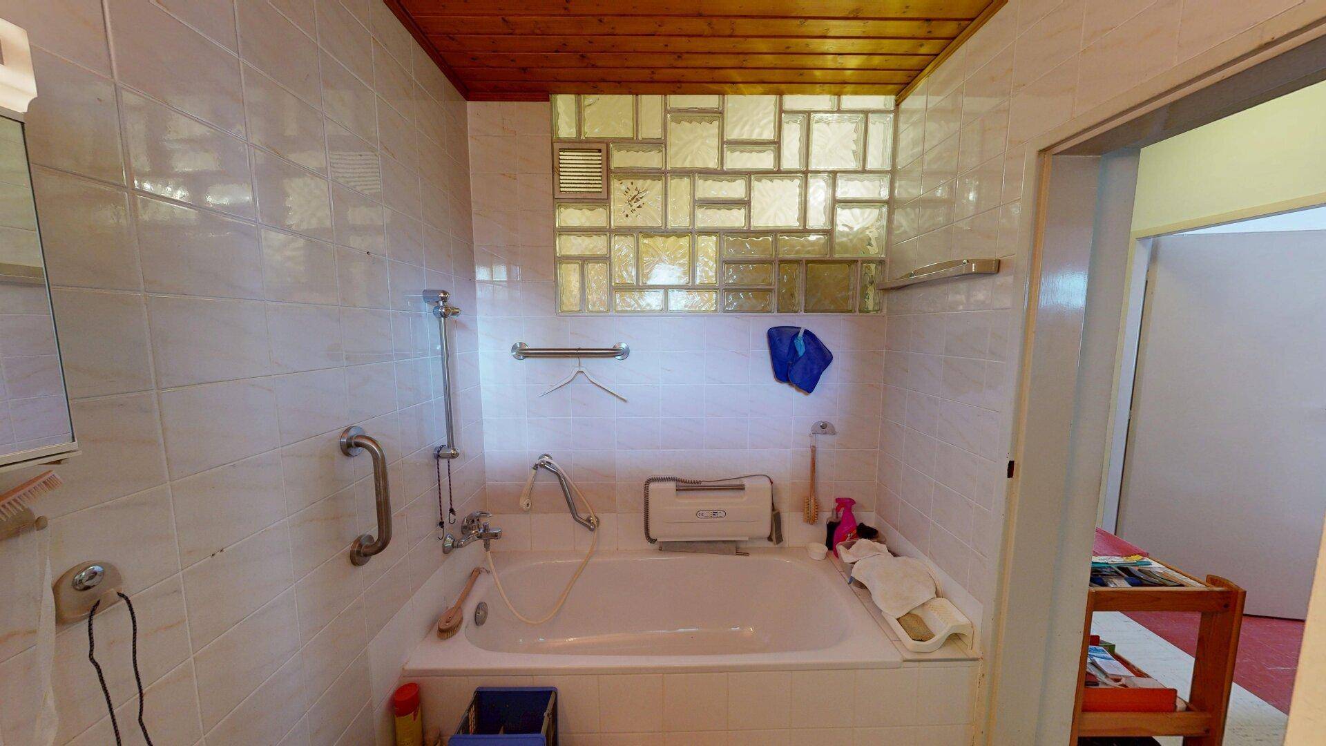 Badezimmer mit Badewanne, Haltegriffen und Glasbausteinfenster, ausgestattet mit Holzdecke und Fliesen.