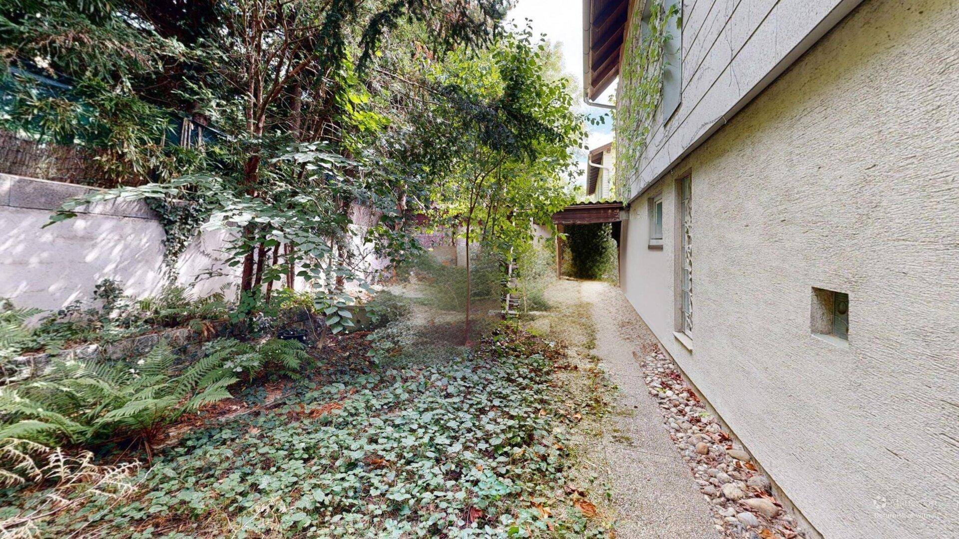 Seitlicher Weg entlang des Hauses, führt zu einem naturbelassenen Gartenbereich mit viel Grün und Bäumen.