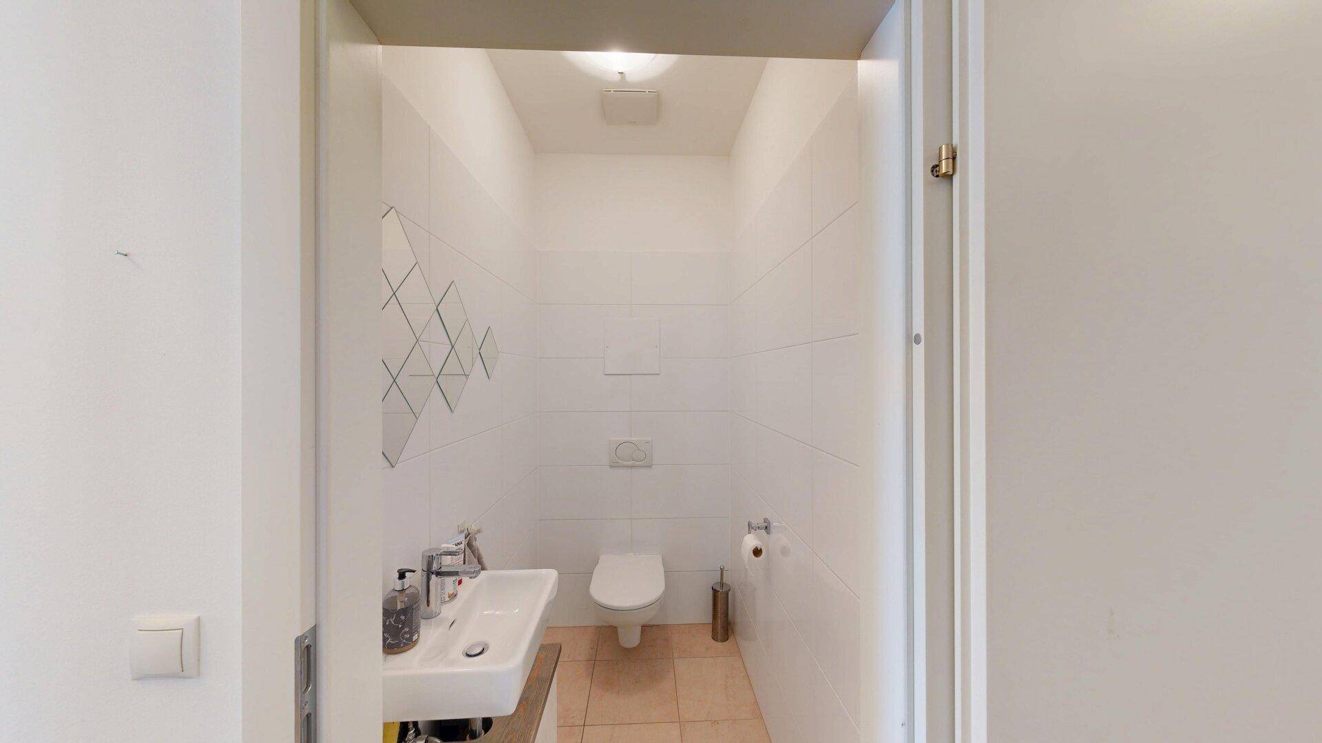 Modernes Gäste-WC mit Wand-WC, kleinem Waschbecken und hellen Fliesen.