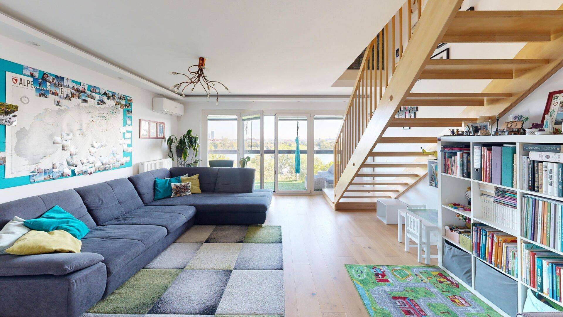 Geräumiger Wohnbereich mit Holztreppe, großem Sofa und Zugang zu einem sonnigen Außenbereich.