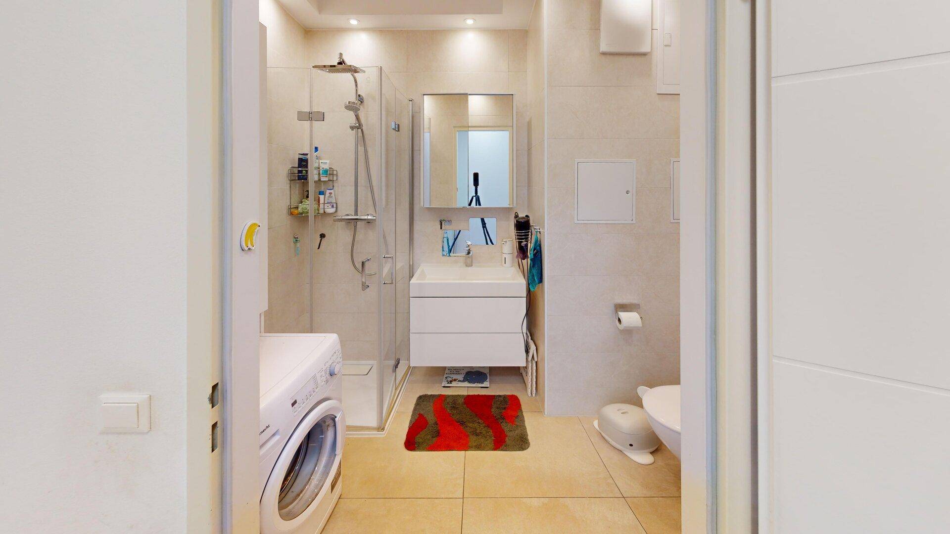 Modernes Badezimmer mit Dusche, Waschmaschine und hellen Fliesen, praktisch und gut beleuchtet.