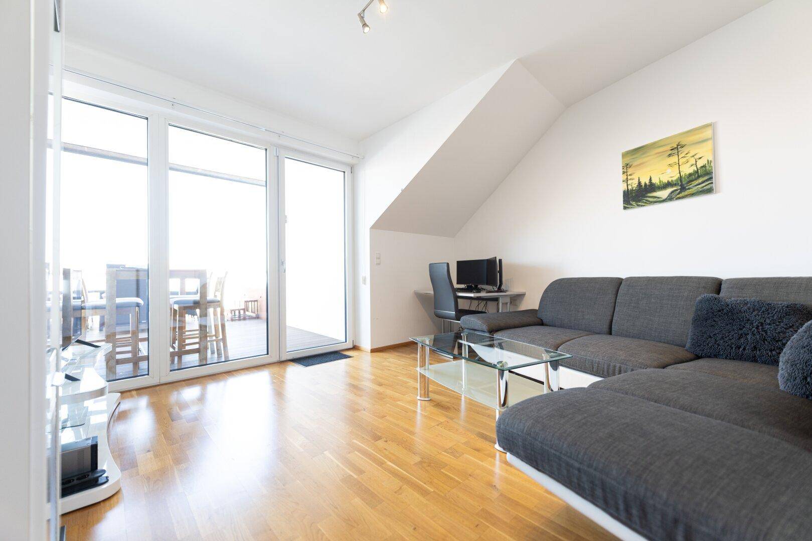 Helles Wohnzimmer mit Sofa, Glastisch und großen Fenstern mit Zugang zum Balkon.