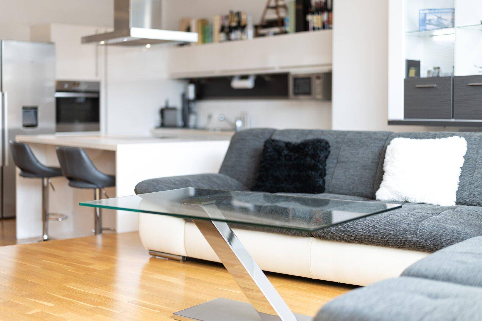 Gemütlicher Wohnbereich mit großem Ecksofa, Glastisch und Blick auf die offene, moderne Küche.
