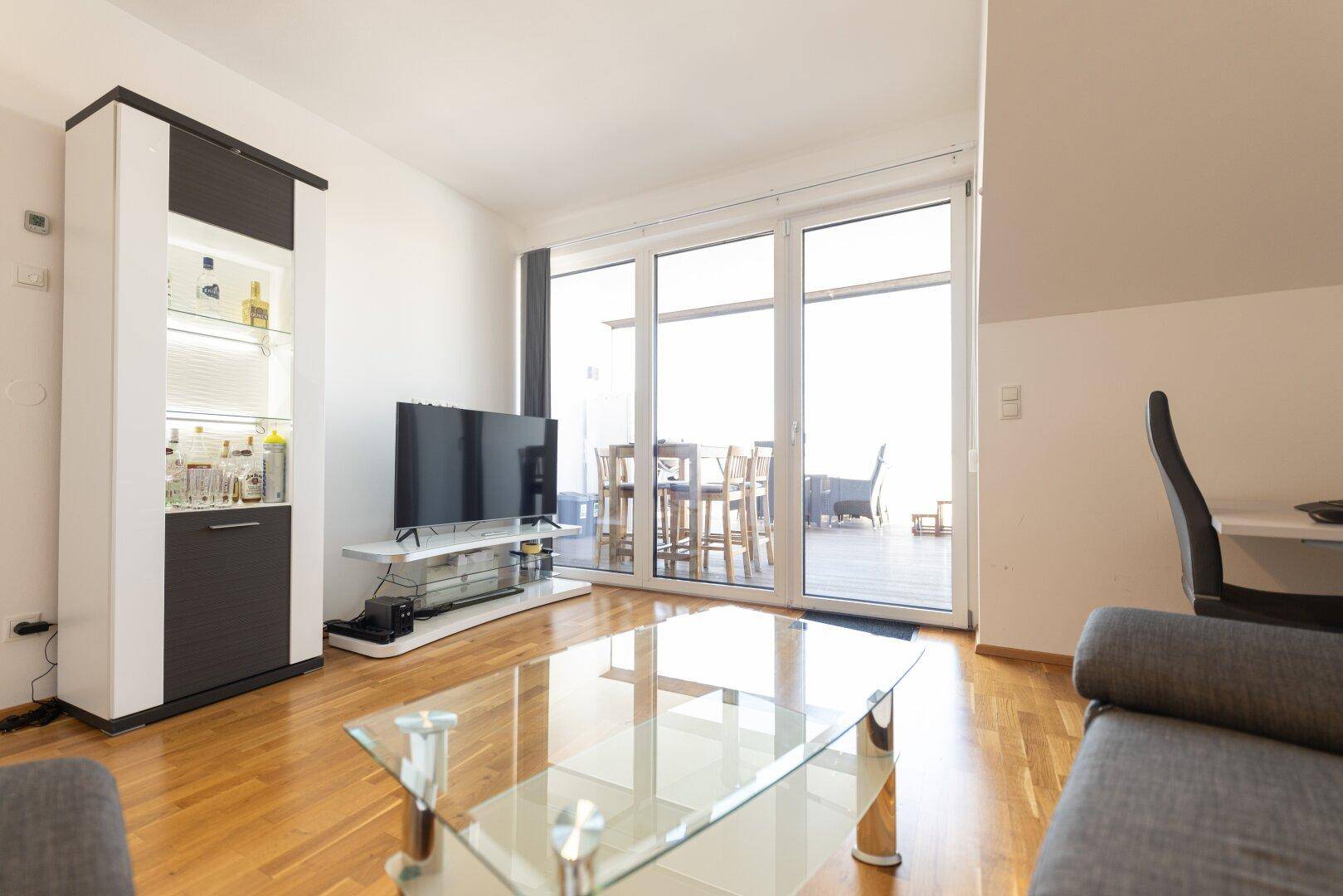 Geräumiges Wohnzimmer mit TV-Möbel, Vitrine und breiter Fensterfront zum Außenbereich.