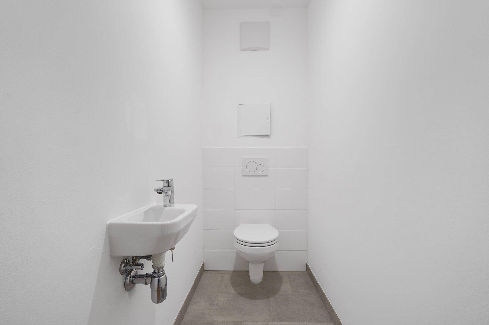 Separates WC mit wandhängender Toilette und kleinem Handwaschbecken, komplett in Weiß gehalten.