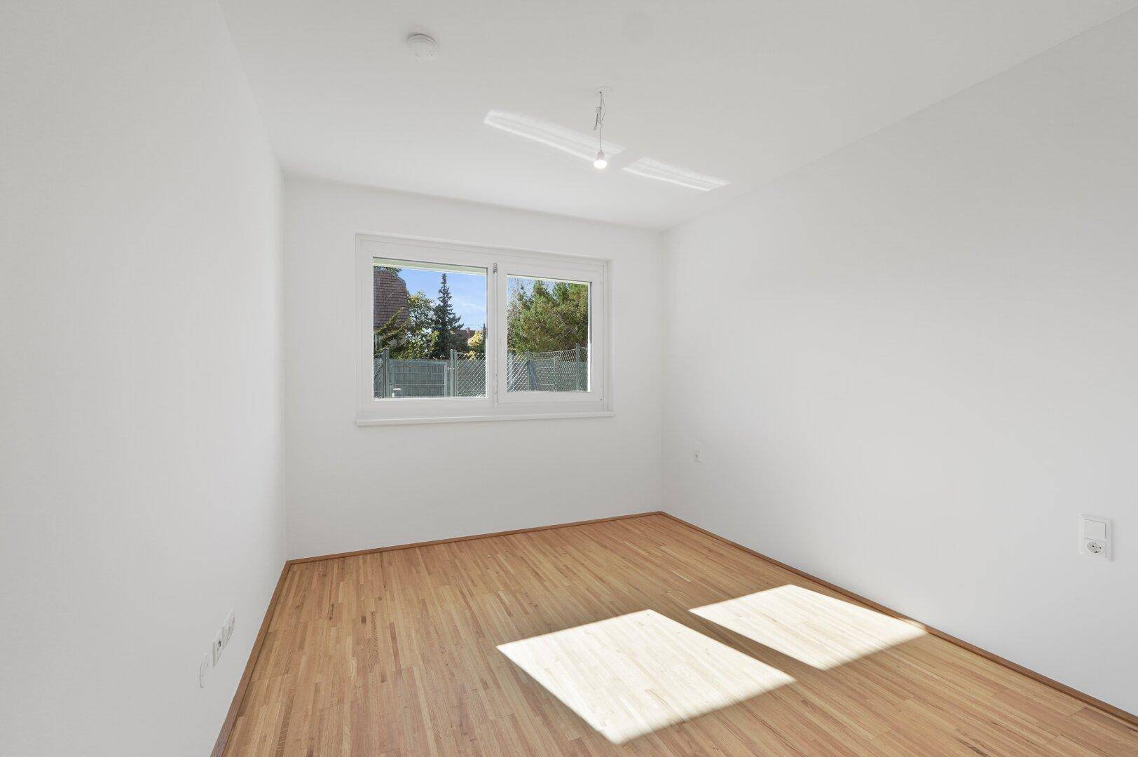 Leeres Schlafzimmer mit Holzfußboden und einem Fenster, das einen schönen Blick ins Grüne bietet.